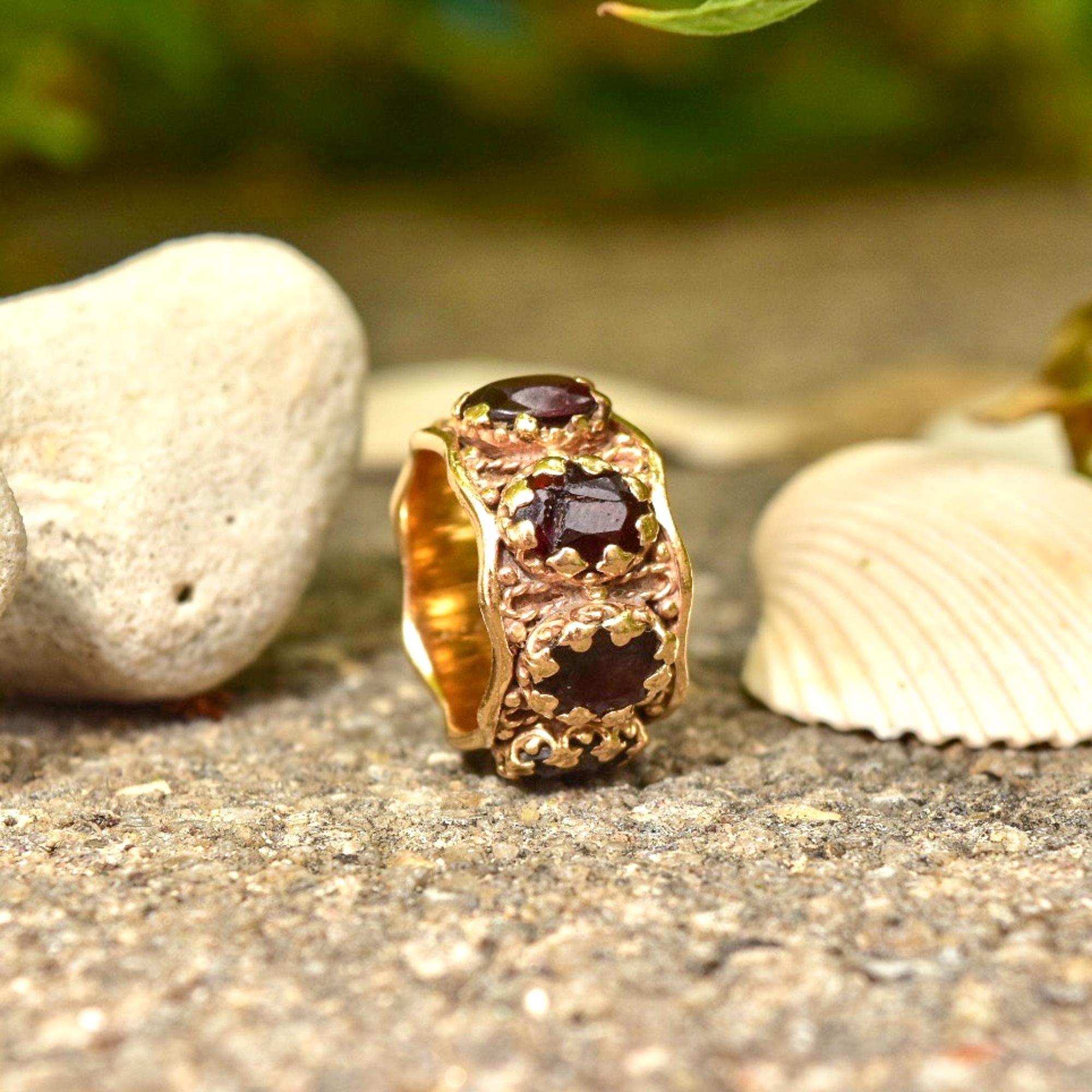 Ein königlicher, breiter 14K-Granat-Ewigkeitsring im etruskischen Stil. Mit einem verzierten 13-mm-Gelbgoldband mit gewellten Kanten, filigranen Details und 7 Granatsteinen im Ovalschliff. Die dunkelroten Edelsteine sitzen sicher in gewellten