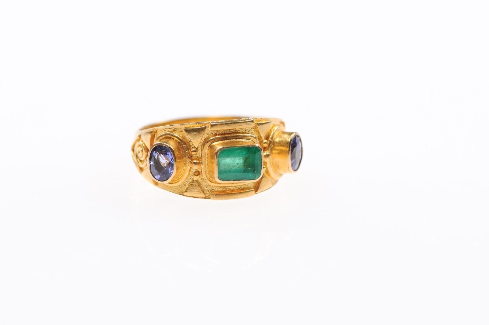 An artisan created gold and gemstone ring, with a beautiful Etruscan style inspired design. Ring features a center emerald, which is flanked by sapphires at either side, set within a 22 karat gold band. Ring size is approximately a ladies 7.5.