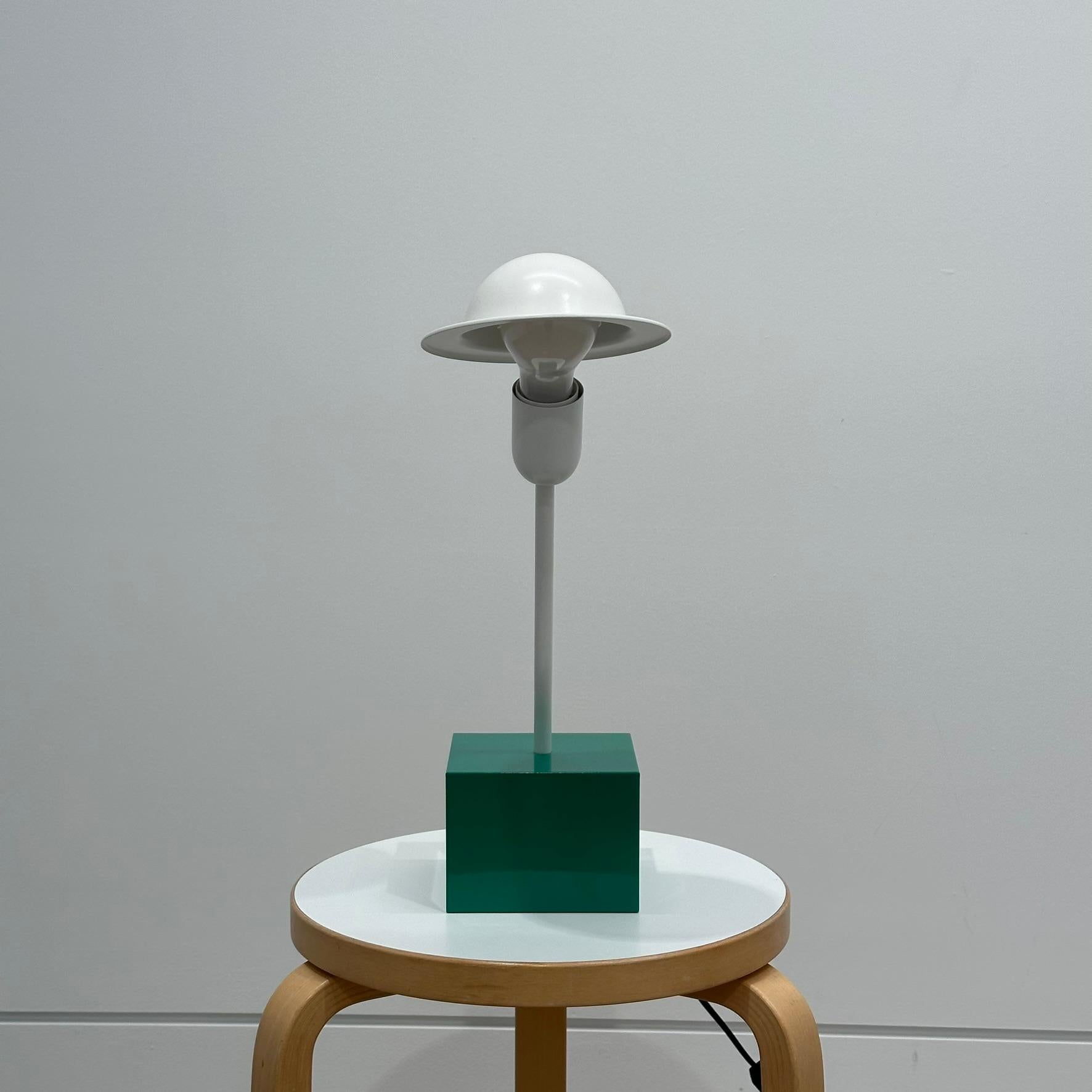 The lamp consists of a relative heavy emerald green cubic base, a white slanted rod and a striking adjustable white shade. This shade is connected to the light bulb with a white lacquered metal clip.