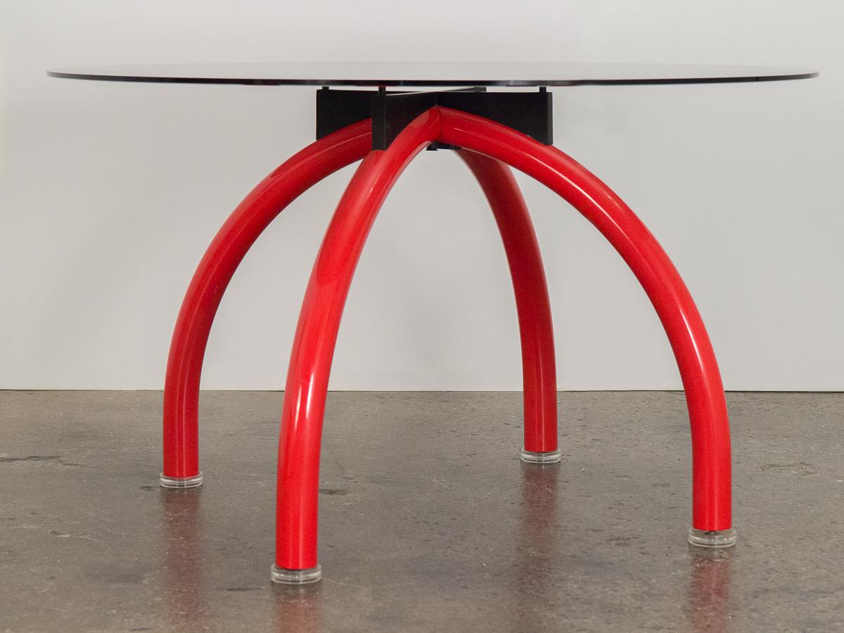 Italian postmodern Spyder dining table designed by Ettore Sottsass for Knoll International. Base is comprised of four curved, tubular steel legs in a bright red enamel finish, balances on adjustable clear Perspex feet. Thick gray glass is secured to