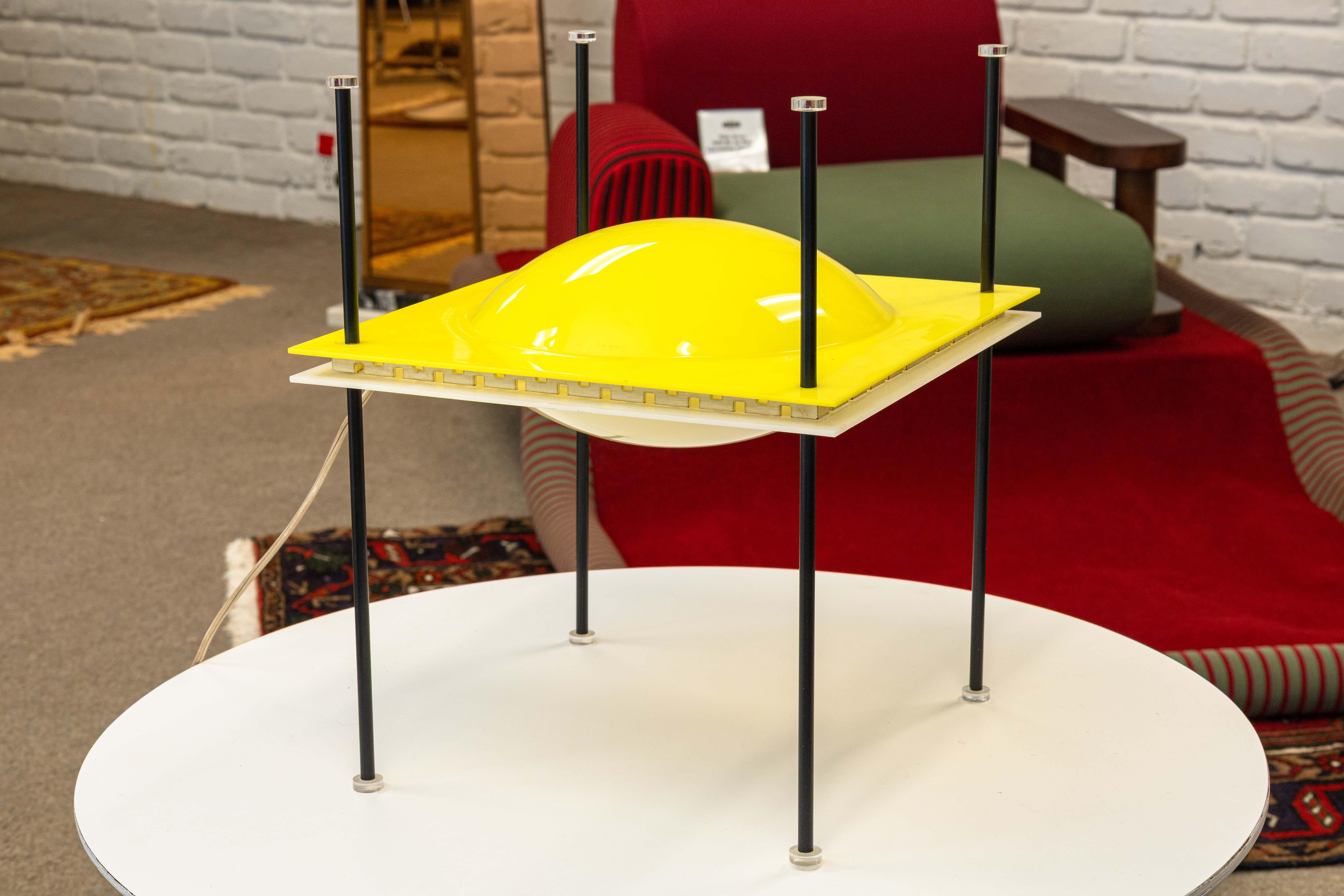  postmodern space age “UFO Lamp” designed by Ettore Sottsass for Arredoluce, Italy 1957. Molded plastic, enameled brass, and lucite. The lamp features a domed yellow diffuser suspended within a square frame supported by four slender black rods. Its