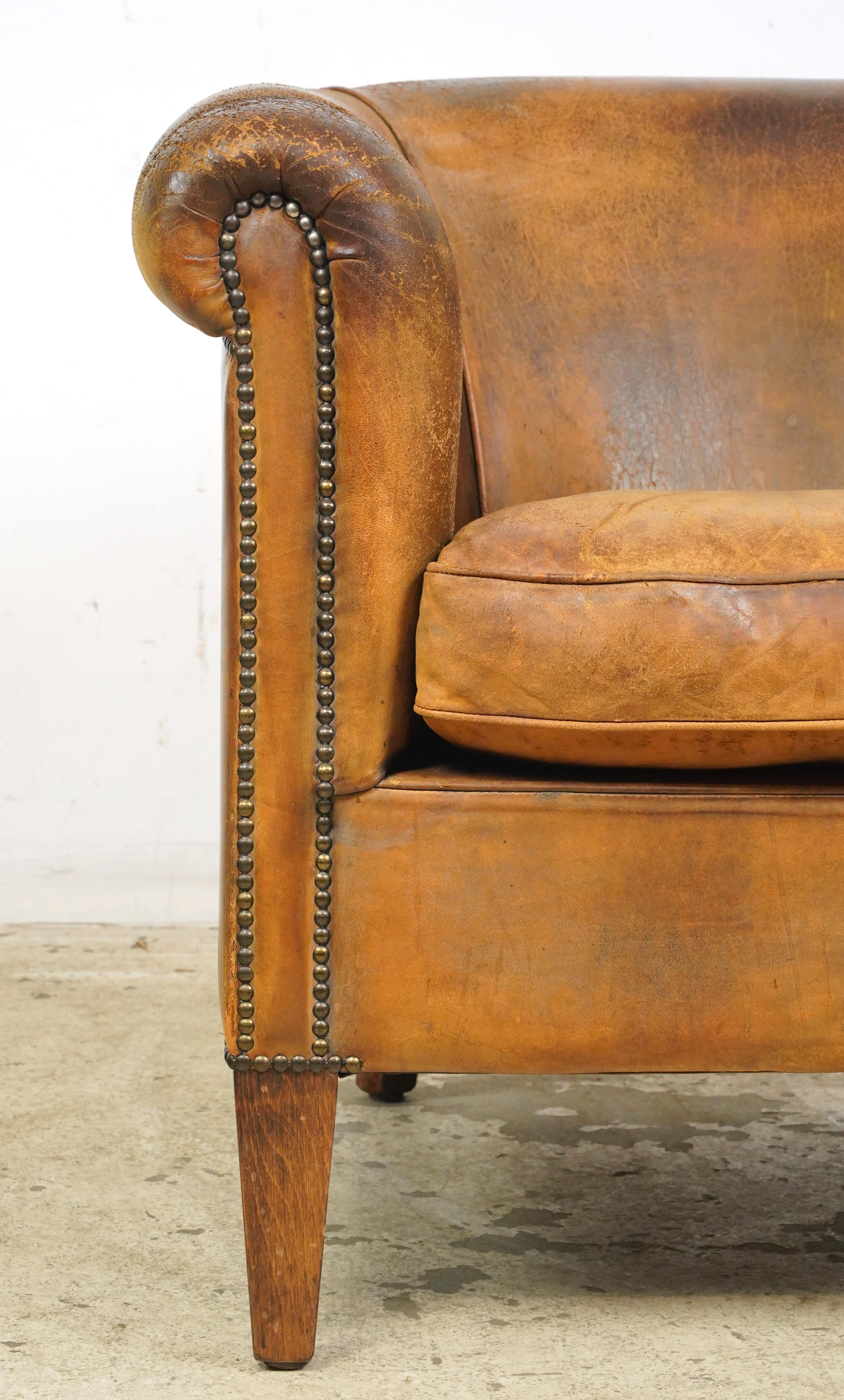 Brown leather studded club chair made in France. It has a round back, wide armrests, a cushioned seat, studded details, and wooden legs. The leather and feet show signs of wear and age. Please refer to the pictures for a full view of the condition.