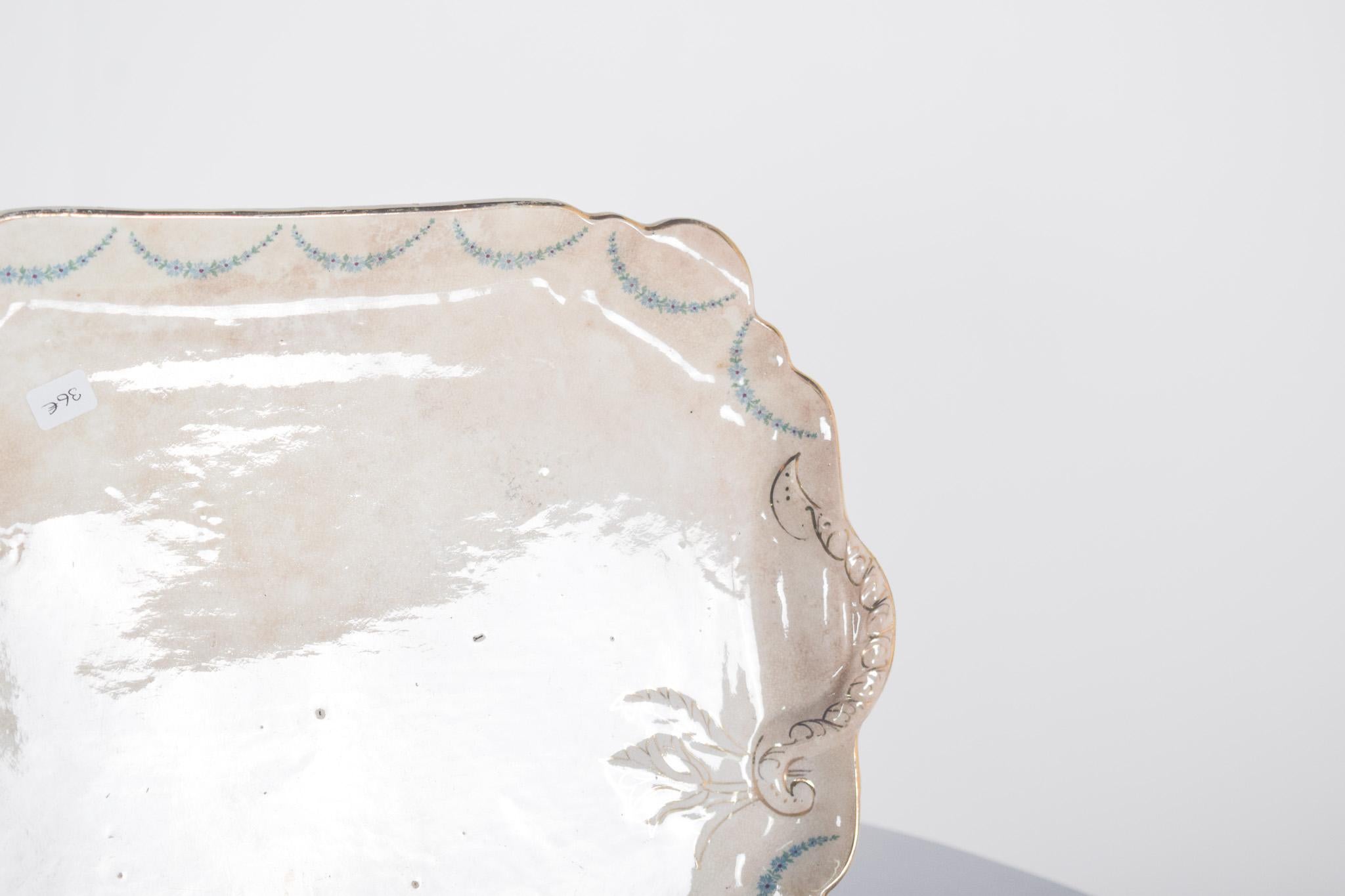 Piatto da portata in ceramica. Rettangolare, con angoli delicatamente arrotondati e tagli, ha una decorazione semplice ed elegante. Lo sfondo ha un effetto leggermente invecchiato, che mostra il passare degli anni.
Con ghirlande di fiori blu che