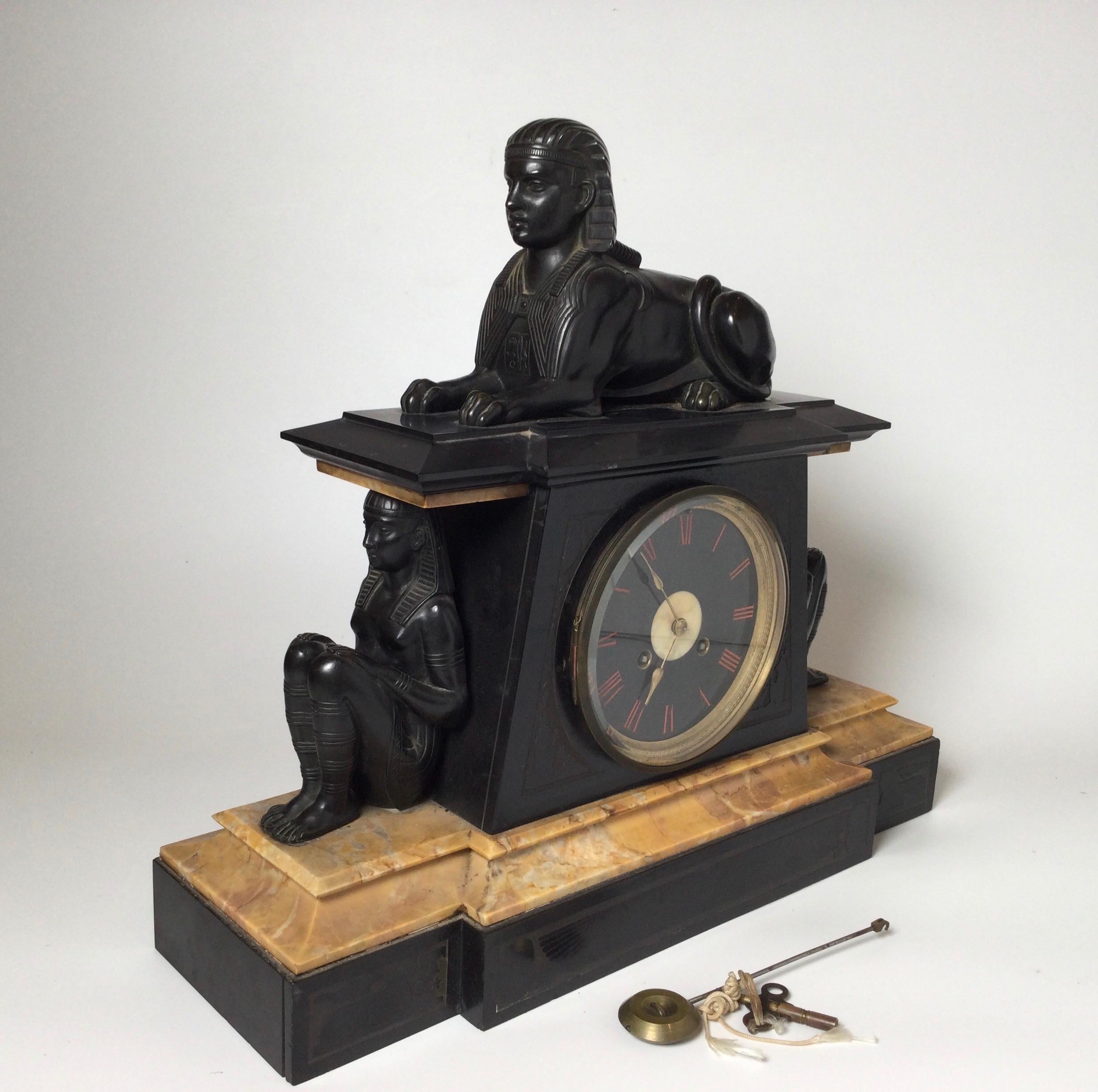 A patinated bronze Egyptian revival figural clock with sphynx top. The bronze figures also flank each side. The Belgian slate with siena marble accents with deep rose colored roman numerals. The clock is running with its original pendulum and key. 8