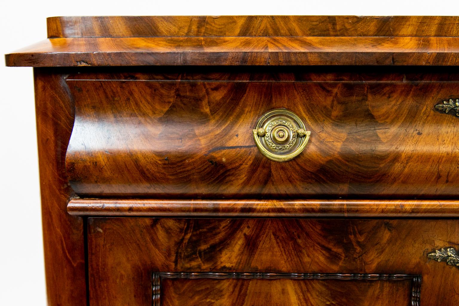 European mahogany Biedermeier commode, the top with bookmatched crotch mahogany veneers above three drawers, one drawer being bombe. The middle drawer has a carved panel and bookmatched veneer in crotch mahogany.
    
