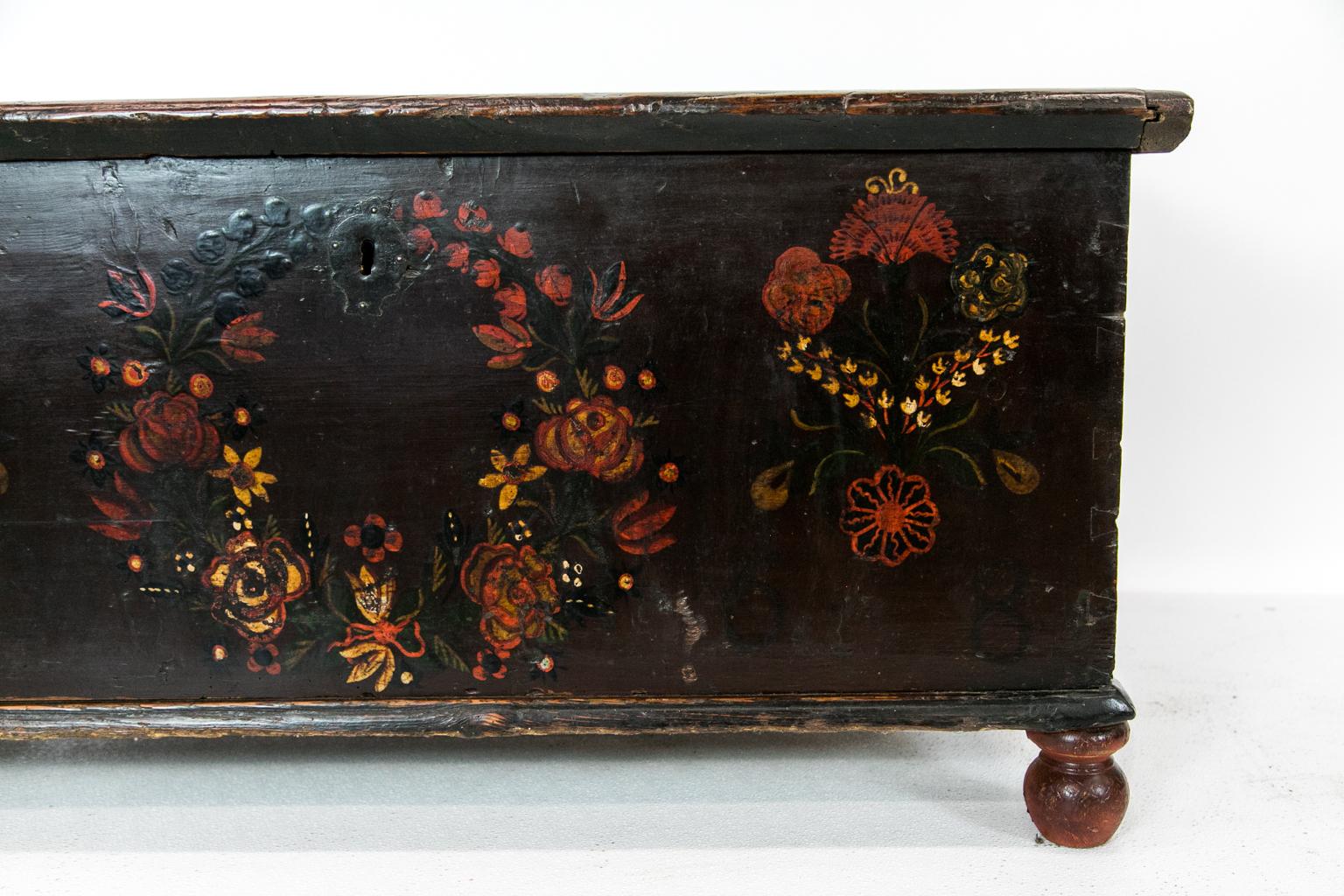 European pine blanket chest is painted brown with a red floral wreath in the center that is flanked on either side with floral bouquets. The interior has the large original steel strapped hinges.
  