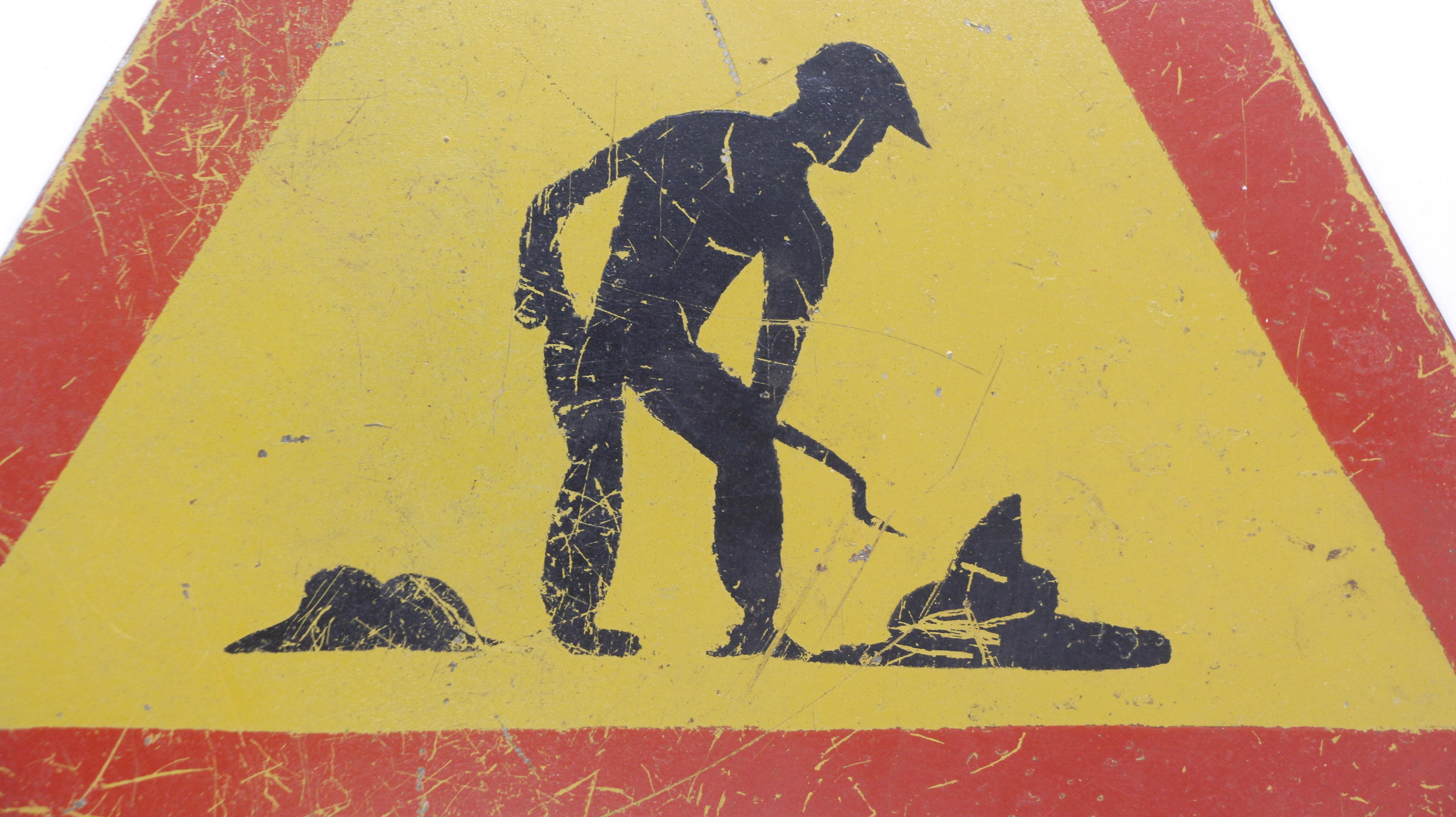 European triangular warning road work sign displaying a worker shoveling icon. It is made of steel in yellow, red, and black colors. There is a pre-drilled hole at the top of the sign for mounting. Good condition with surface distressed wear from