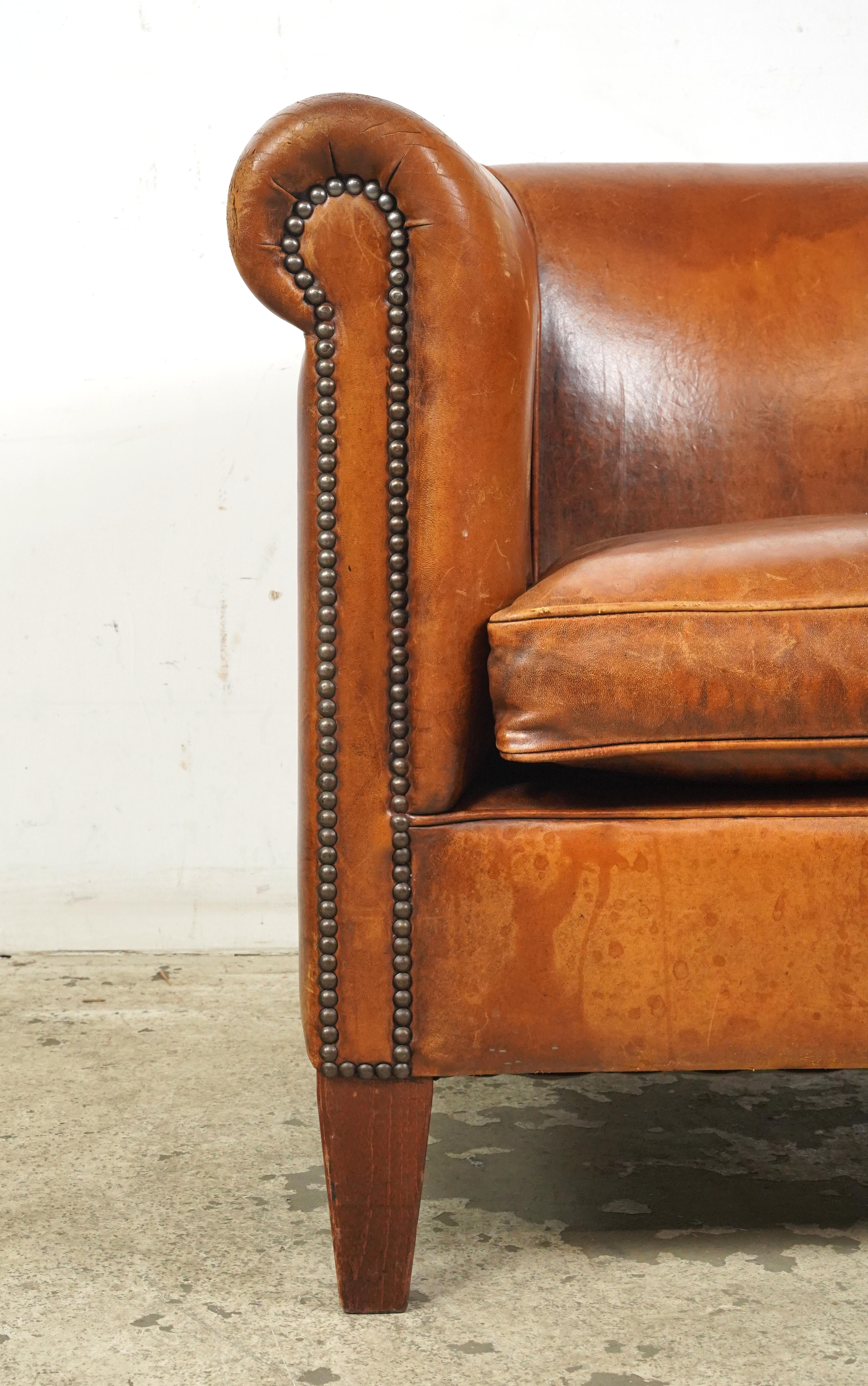 Brown leather studded club chair made in France. It has a round back and arms, a cushioned seat, studded details, and wooden legs. The leather and feet show signs of wear and age. Please refer to the pictures for a full view of the condition. One