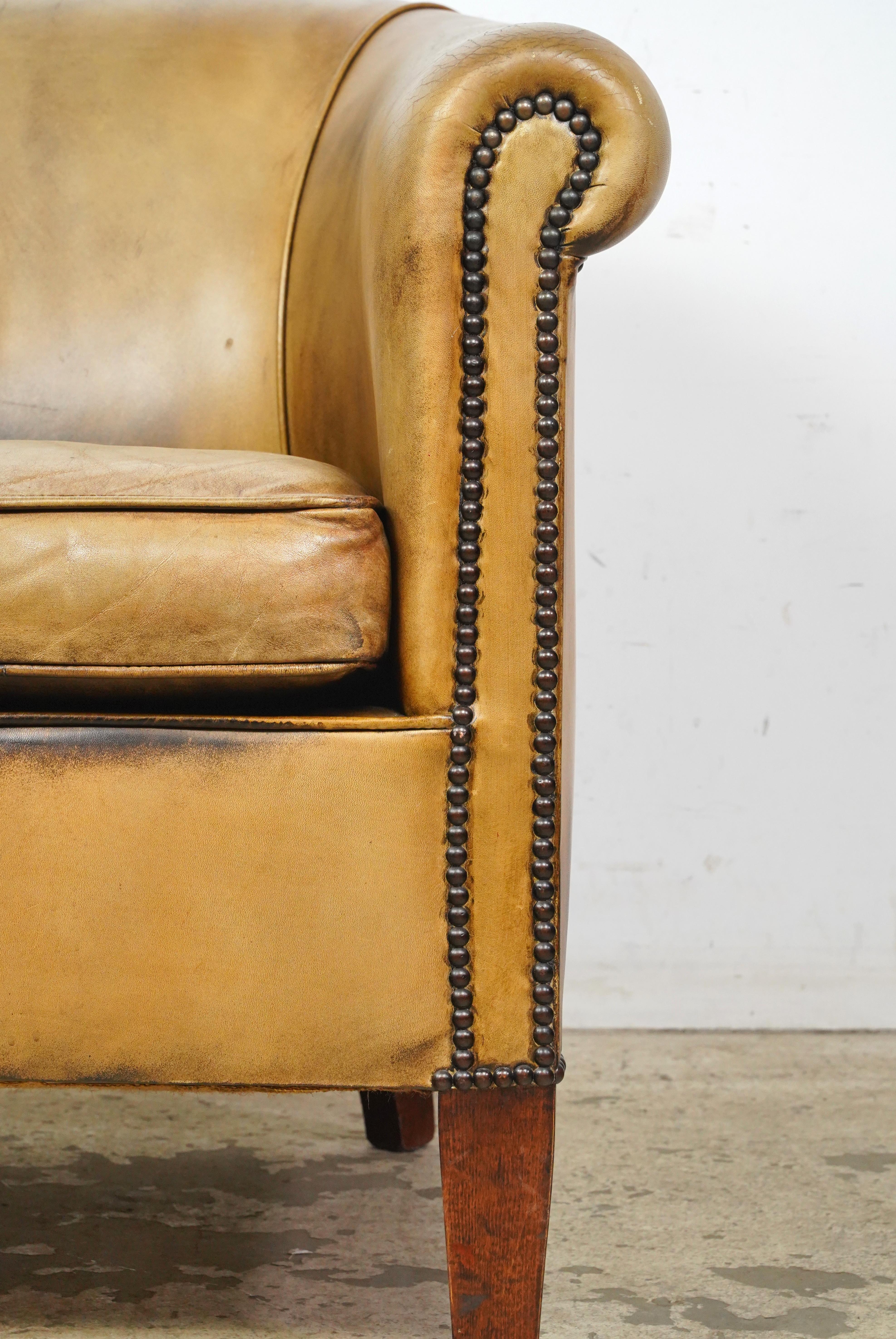Tan leather studded club chair made in France. It has a round back, wide armrests, a deep seat, studded details, and wooden legs. The leather and feet show minor signs of wear and age. Please refer to the pictures for a full view of the condition.
