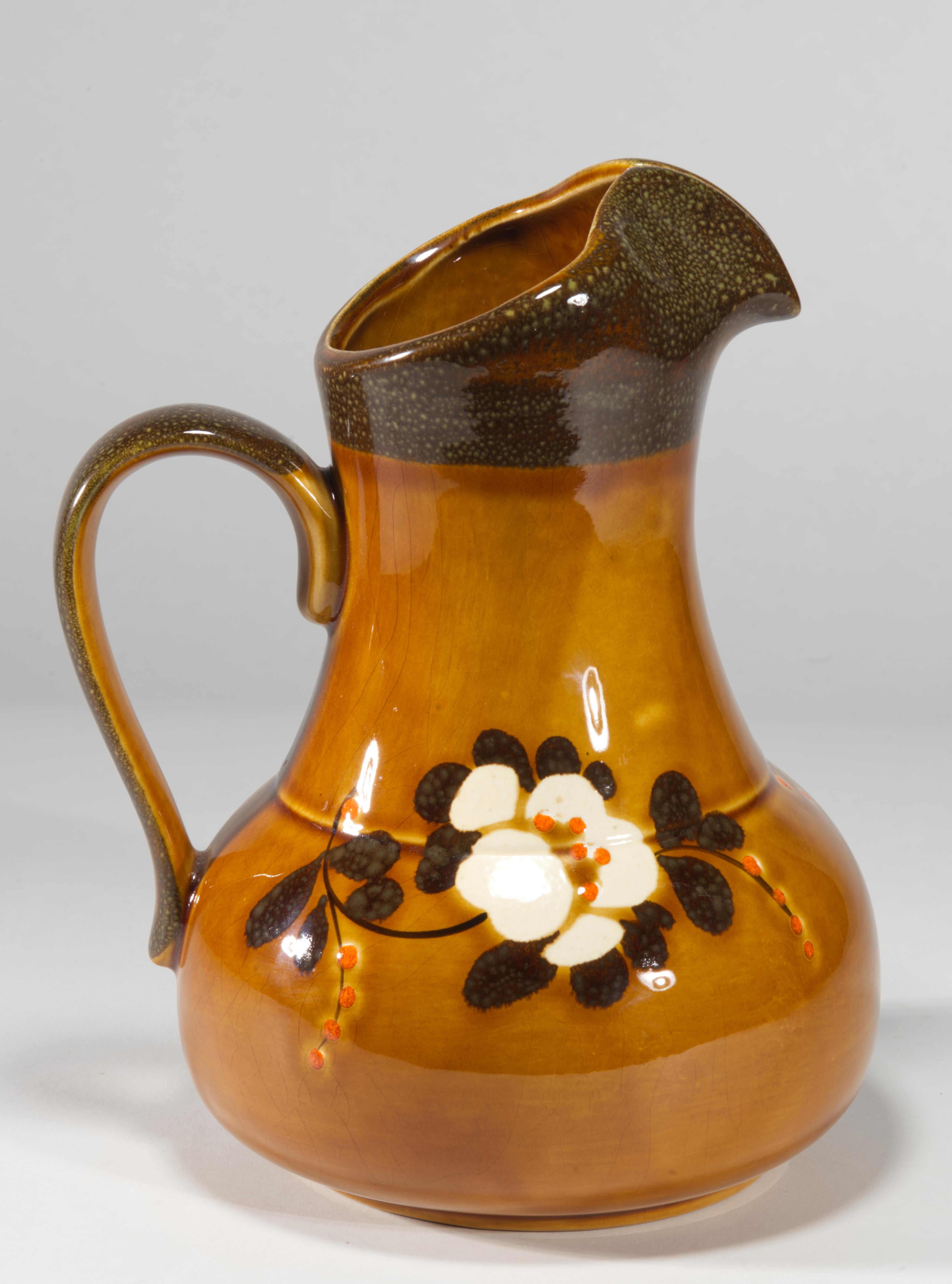 Il vaso scultoreo a forma di brocca presenta una forma morbida e arrotondata con bordo e manico curvi; è dipinto a mano con motivi floreali basati sulla Folk Art e fortemente influenzati dall'Art Déco. I colori bianco, blu cobalto e arancione dei