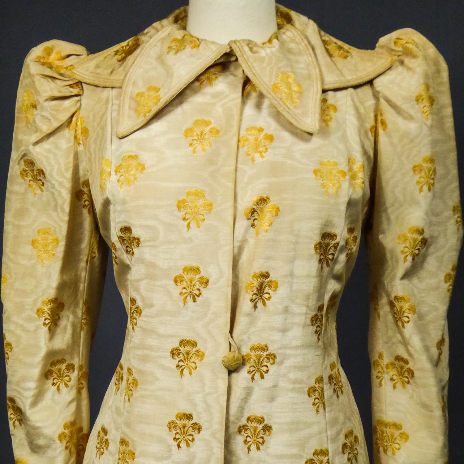 Circa 1935/1940
England

Amazing british evening or interior coat in straw-yellow silk moire brocaded in gold and dating from the years 1935/1940. Rich silk façonné with golden trimmed bunches recalling a stylized fleur-de-lis on a straw-yellow silk