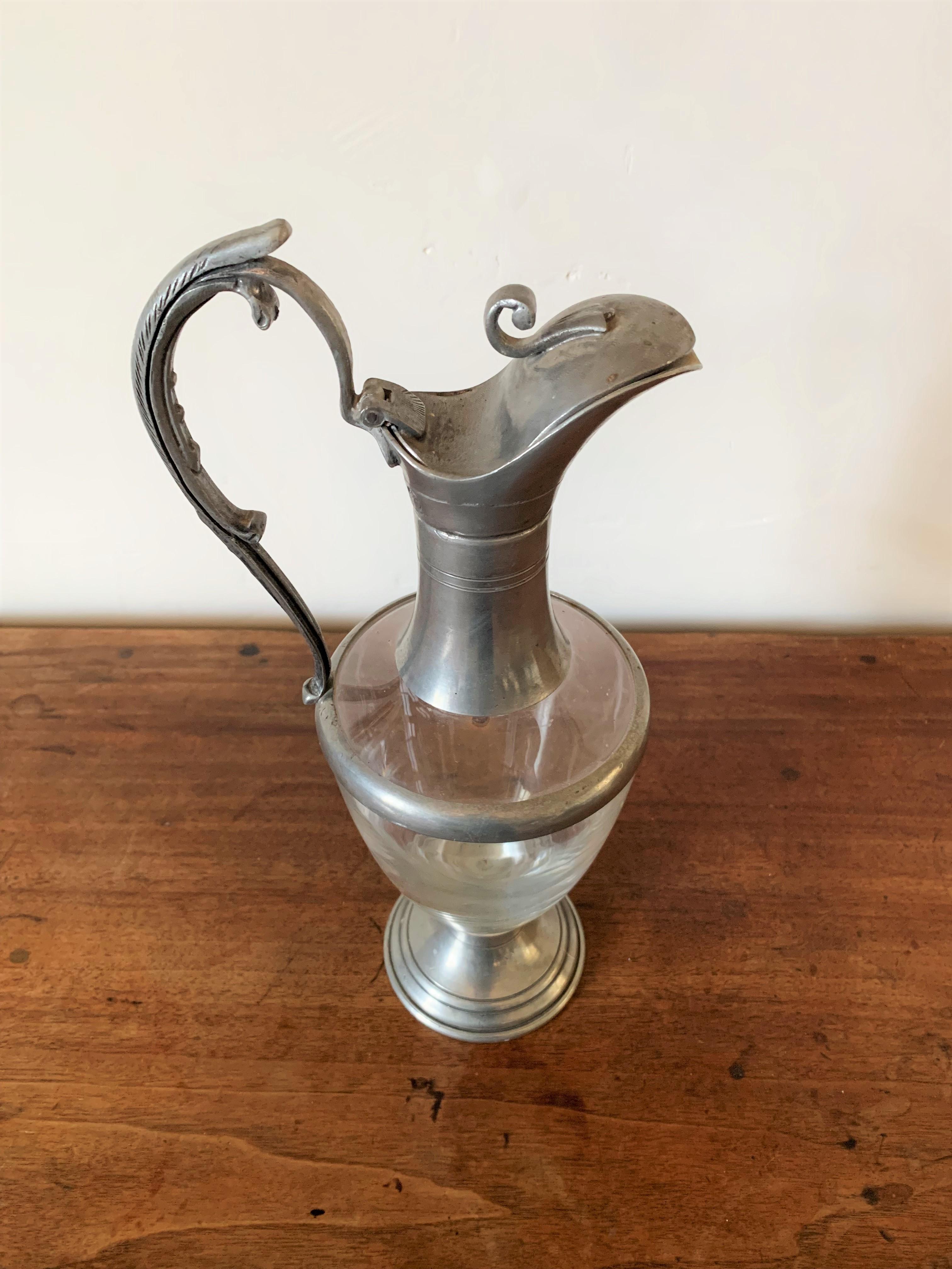 Very nice ewer, carafe from the early 20th century. The body is made of smooth glass while the foot, handle and spout are made of pewter, a very rare element and one of the oldest known at least since antiquity. The handle is graceful with a vegetal
