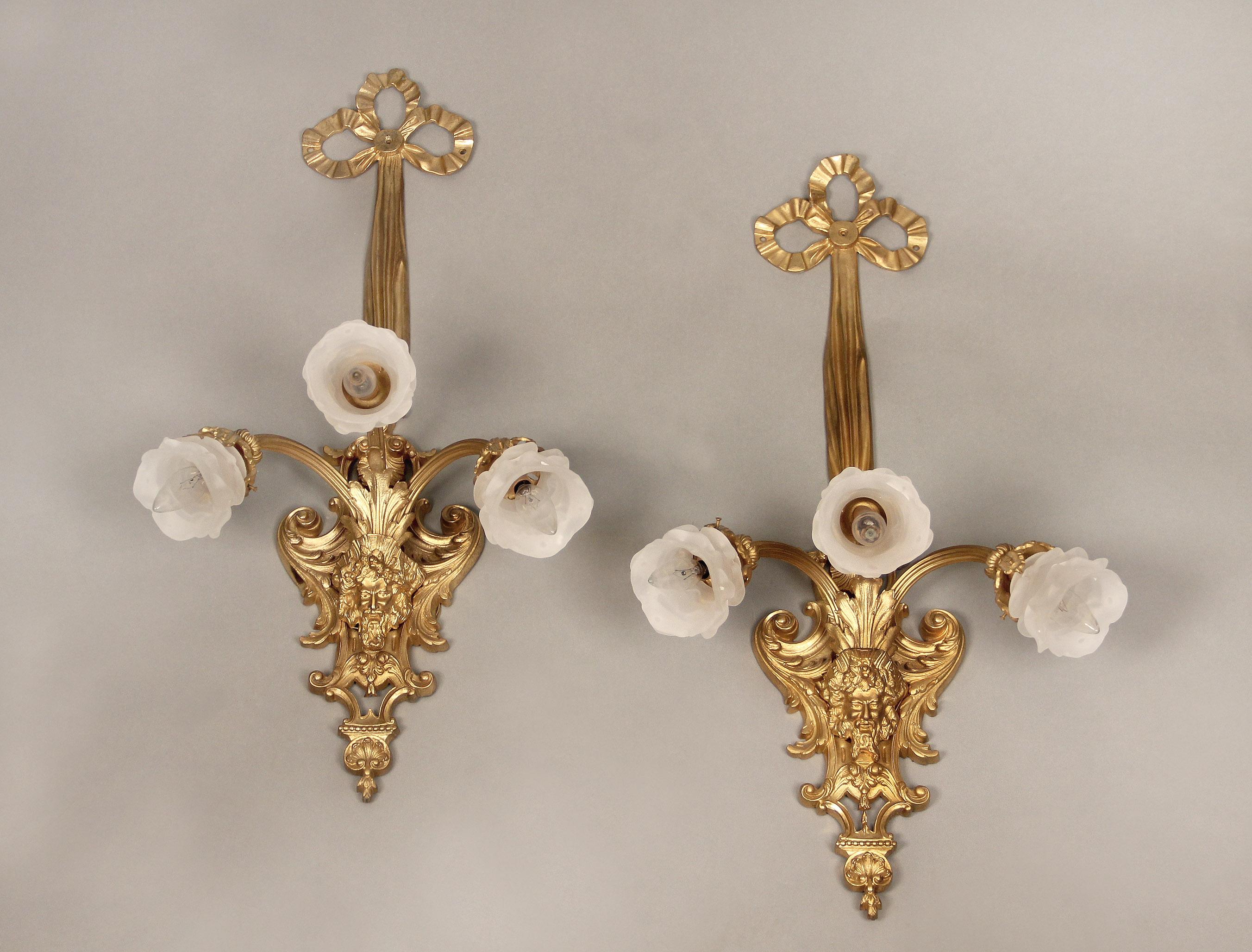 An excellent pair of late 19th century gilt bronze three-light sconces

The center of the backplate depicting a bearded man, the top with a tied ribbon. Three arms with rose shades.