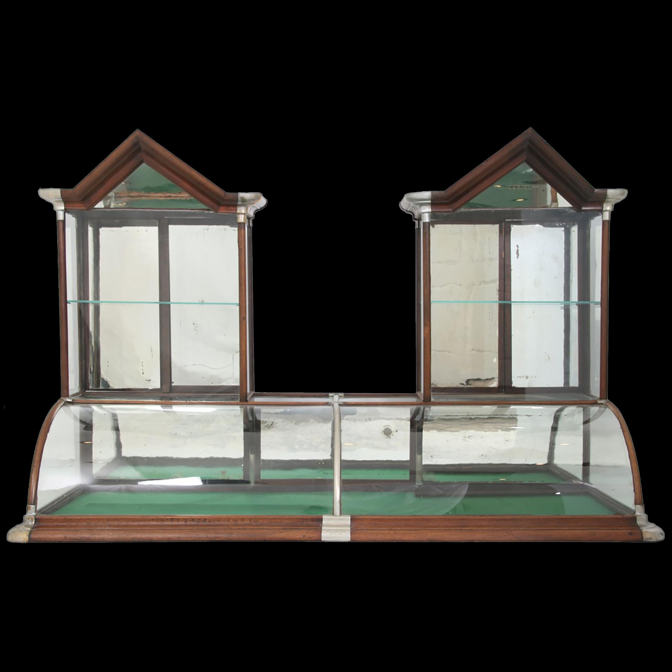 Manufactured by Excelsior Showcase Works, has a curved glass front with walnut trim and nickel-silver mounts and corner trim, has 2 towers on the top with overhanging pointed pediments with mirrored peaks and mirrored sliding doors in the rear.