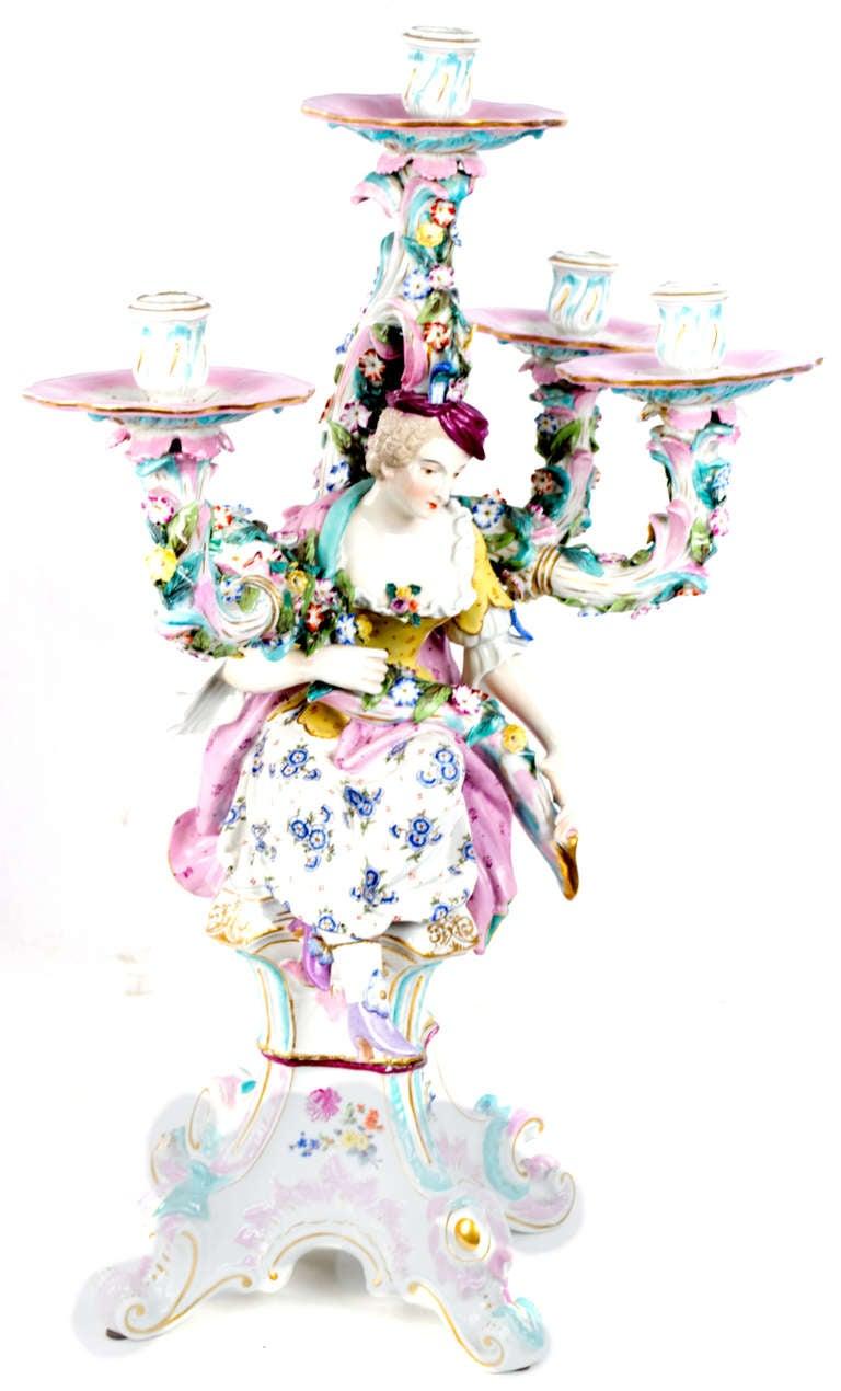 The center of the porcelain candelabra features the figures of a woman, inspired by 18th century German courtly dress, decorated in bright colors and patters. The auricular arms of the candelabra rise above her head like a tree, festooned with