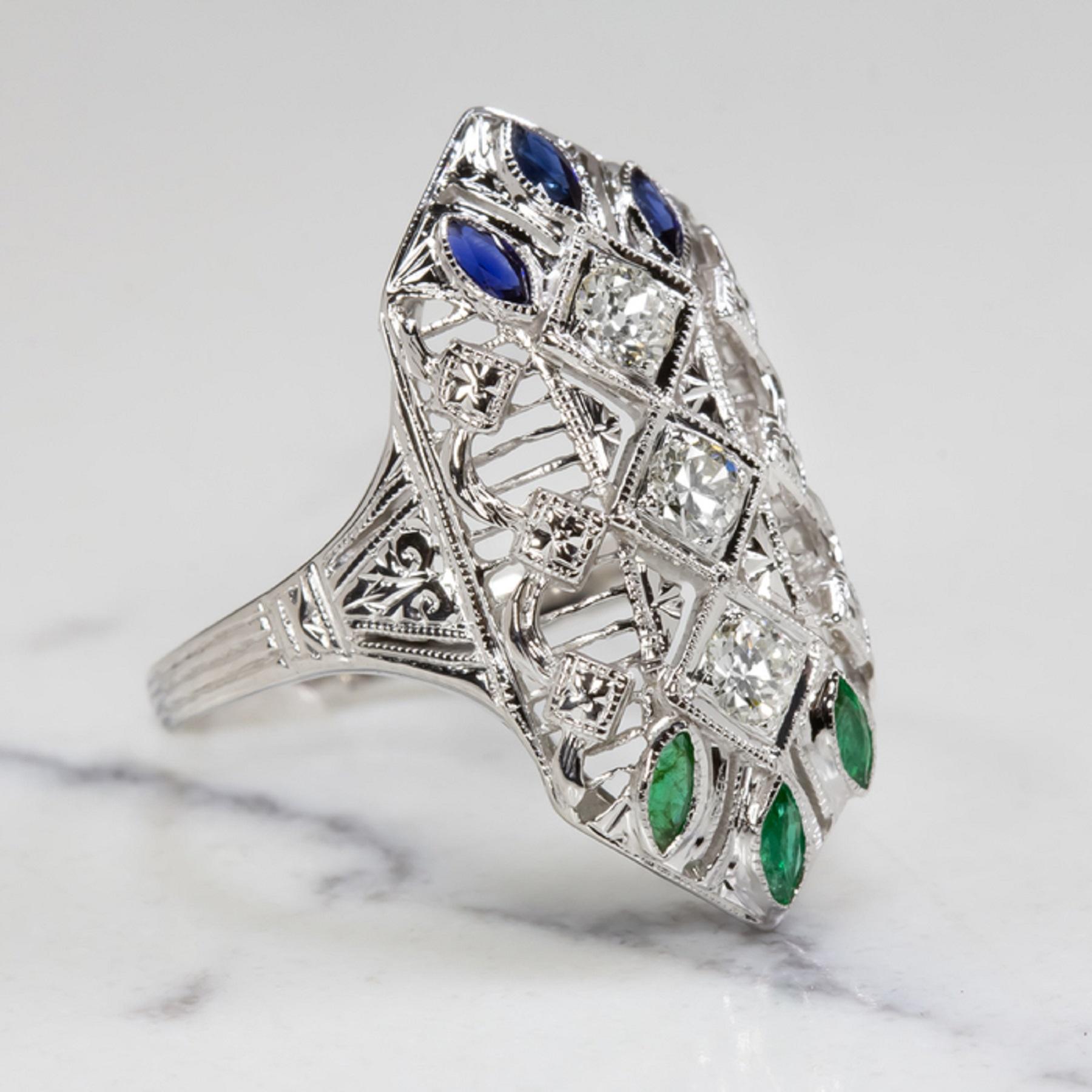 This gorgeous diamond, emerald, and sapphire studded cocktail ring is elegantly designed with an artful structure and was masterfully crafted by hand a lifetime in the past during the illustrious Art Deco era. 

The ring features a trio of old