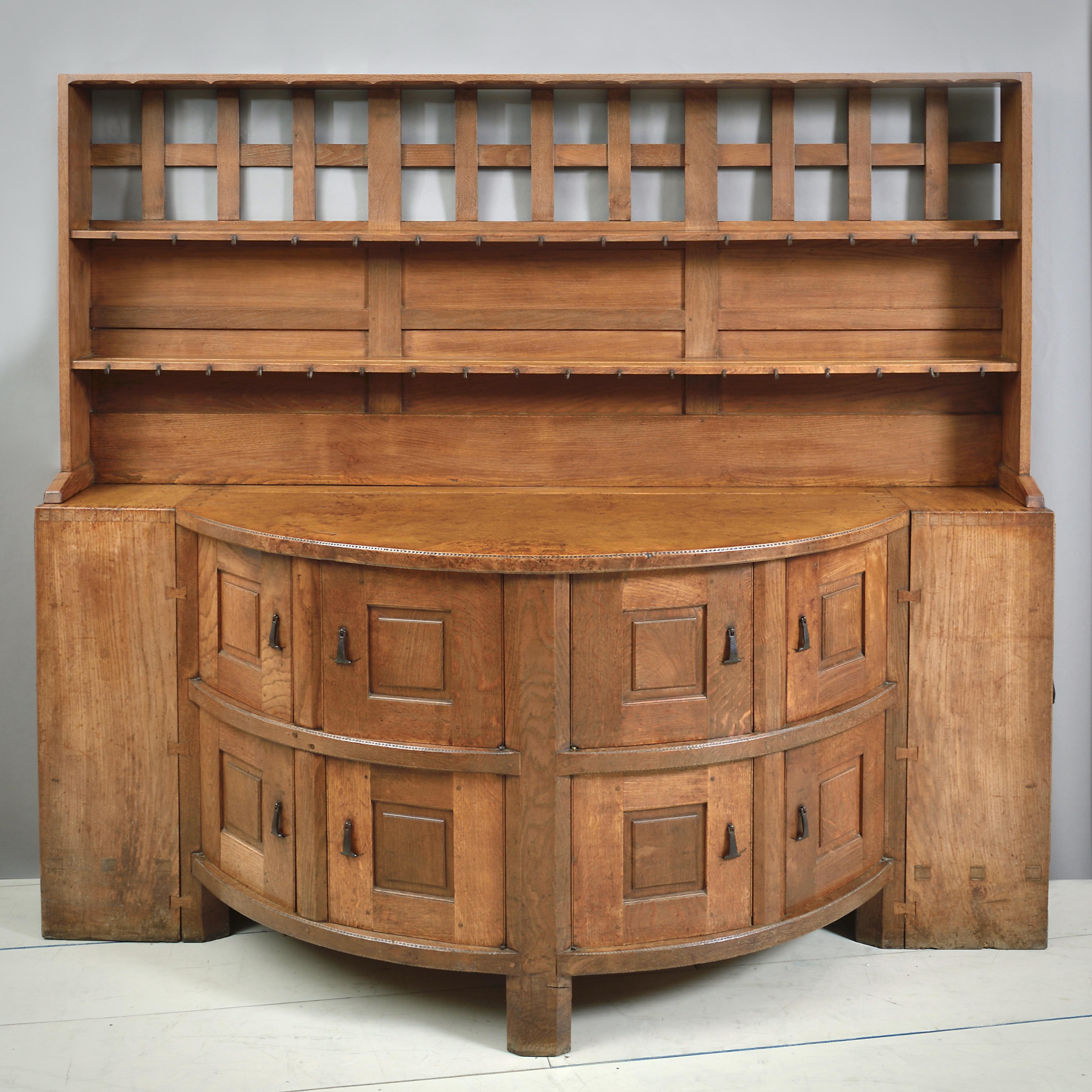 AN EXCEPTIONAL ARTS & CRAFTS OAK DRESSER BY SIDNEY BARNSLEY, CIRCA 1900.

The plate rack with grooved shelves fitted with iron cup hooks, the bow-front base with eight panelled doors, and with a door to each side enclosing three shelves. With