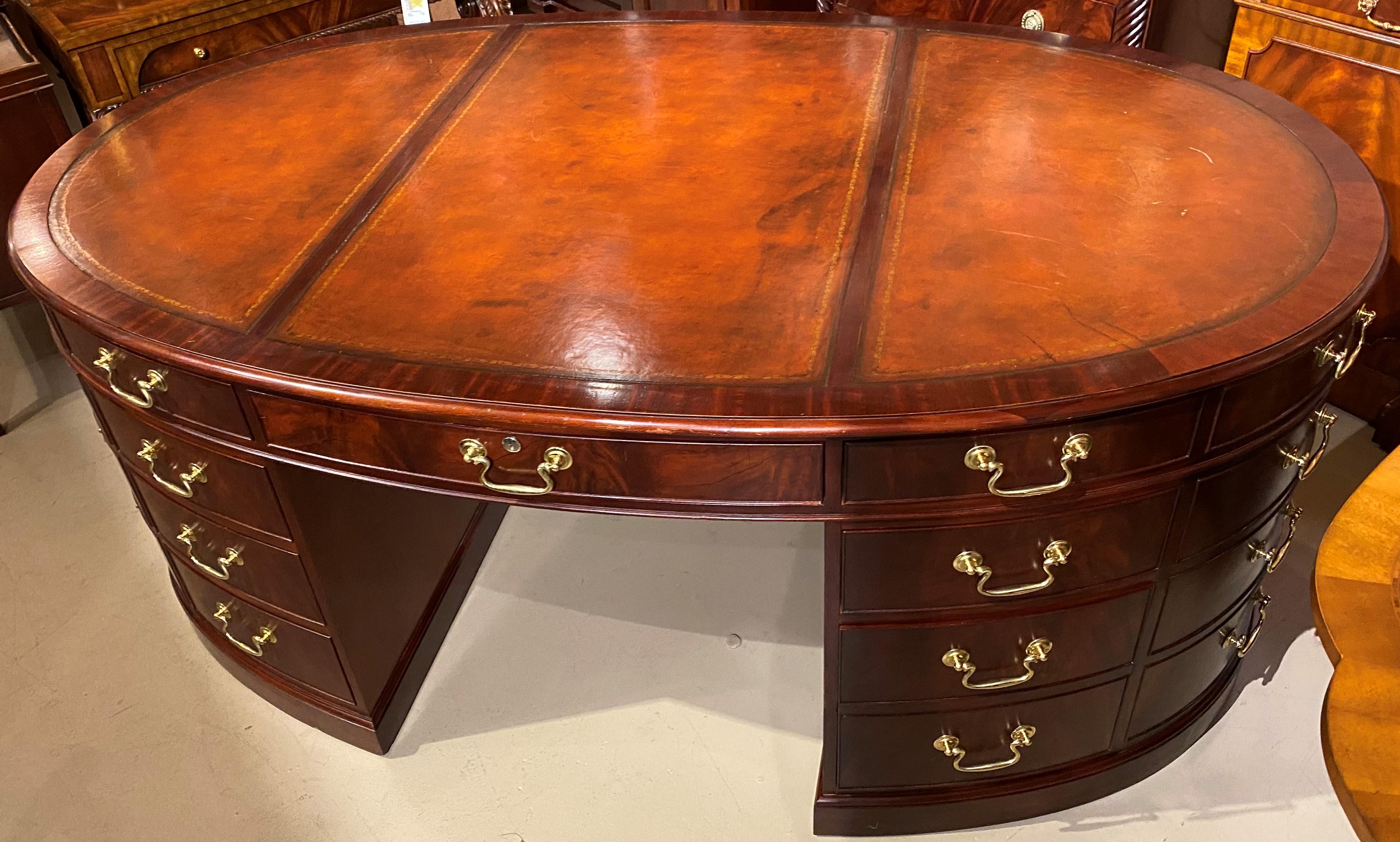 A fine quality oval mahogany partners desk by Baker Furniture Company with three-section brown leather top with gilt tooled border, one side with eight cockbeaded drawers, including a deep file drawer on the right side, and the other side with a
