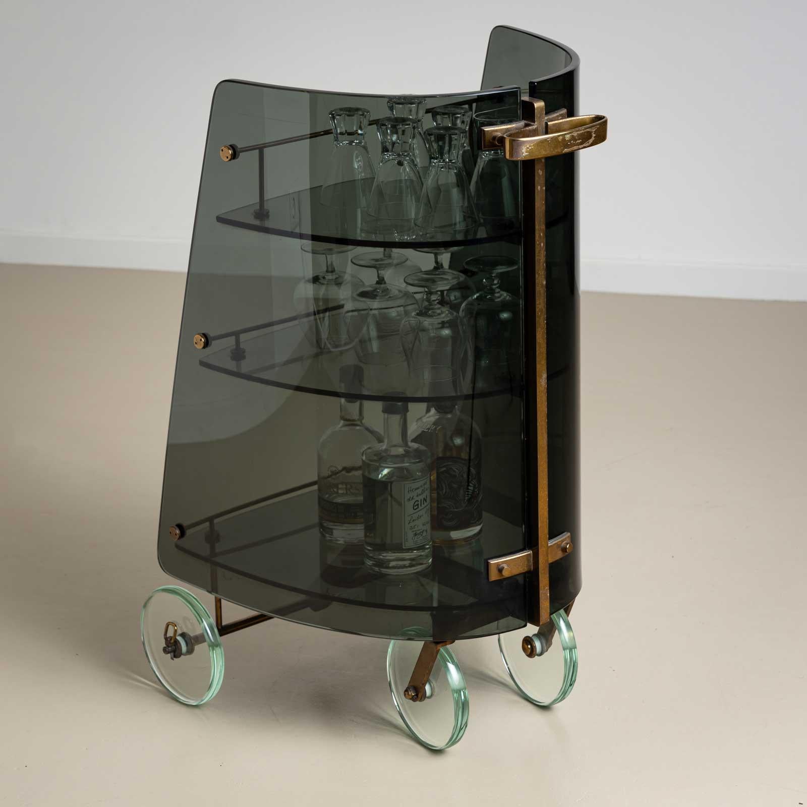 An Italian mid-century bar trolley crafted from smoked glass with refined brass fittings. The trolley rests on four wheels, also made of glass, enhancing its sleek and sophisticated design. It features three shelves that gradually increase in size