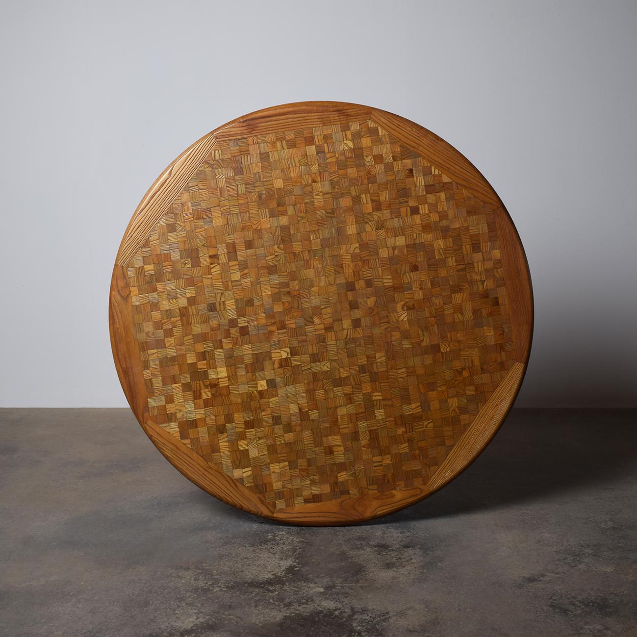 A Stunning mid-century modern mosaic oak table. The design features a circular tabletop with a mosaic oak veneer, showcasing a rich patchwork of natural wood grain patterns. The surface is subtly glossy, highlighting the craftsmanship and the