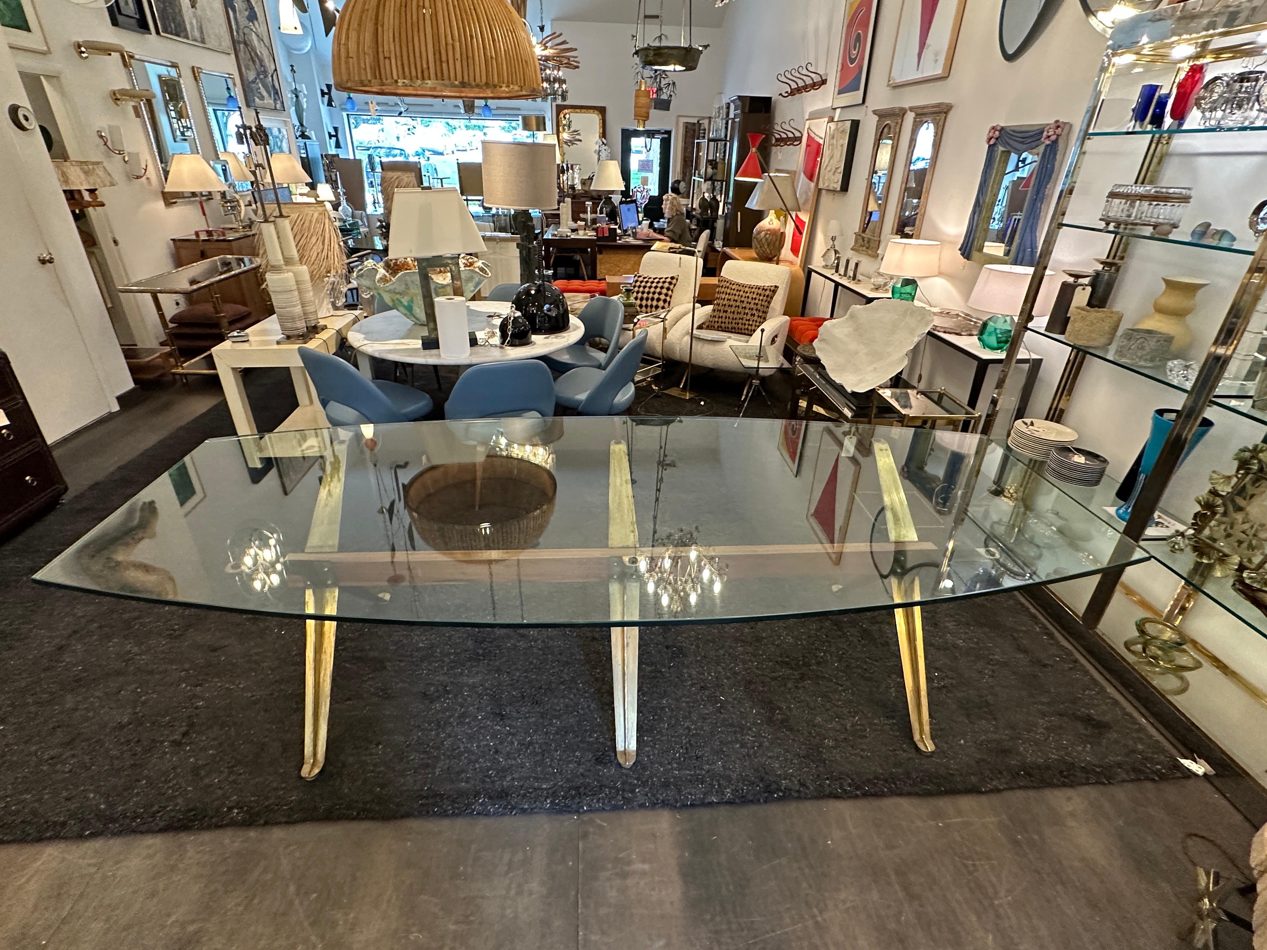 Cette table surdimensionnée très architecturale, avec ses trois pieds en X en laiton et sa poutre en noyer, est parfaite pour une salle à manger,  galerie, salle de conférence, bibliothèque.  Le plateau en verre épais est ovalisé avec une coupe
