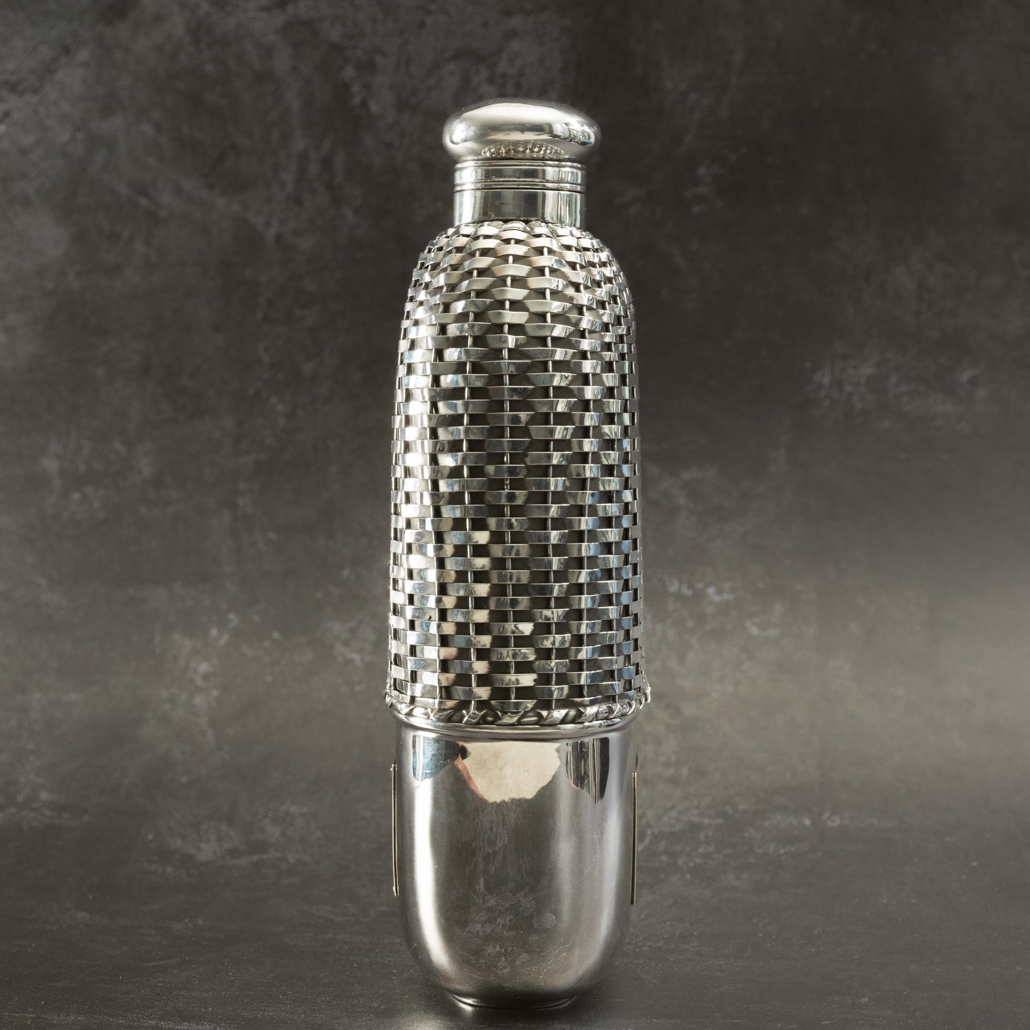 Unusually large silver and glass flask with the silver on the top half of the body woven to resemble wicker basket work. Has cork lined stopper on a hinged bayonet closer, stamped Sterling. A silver removable cup covers the lower half with gold