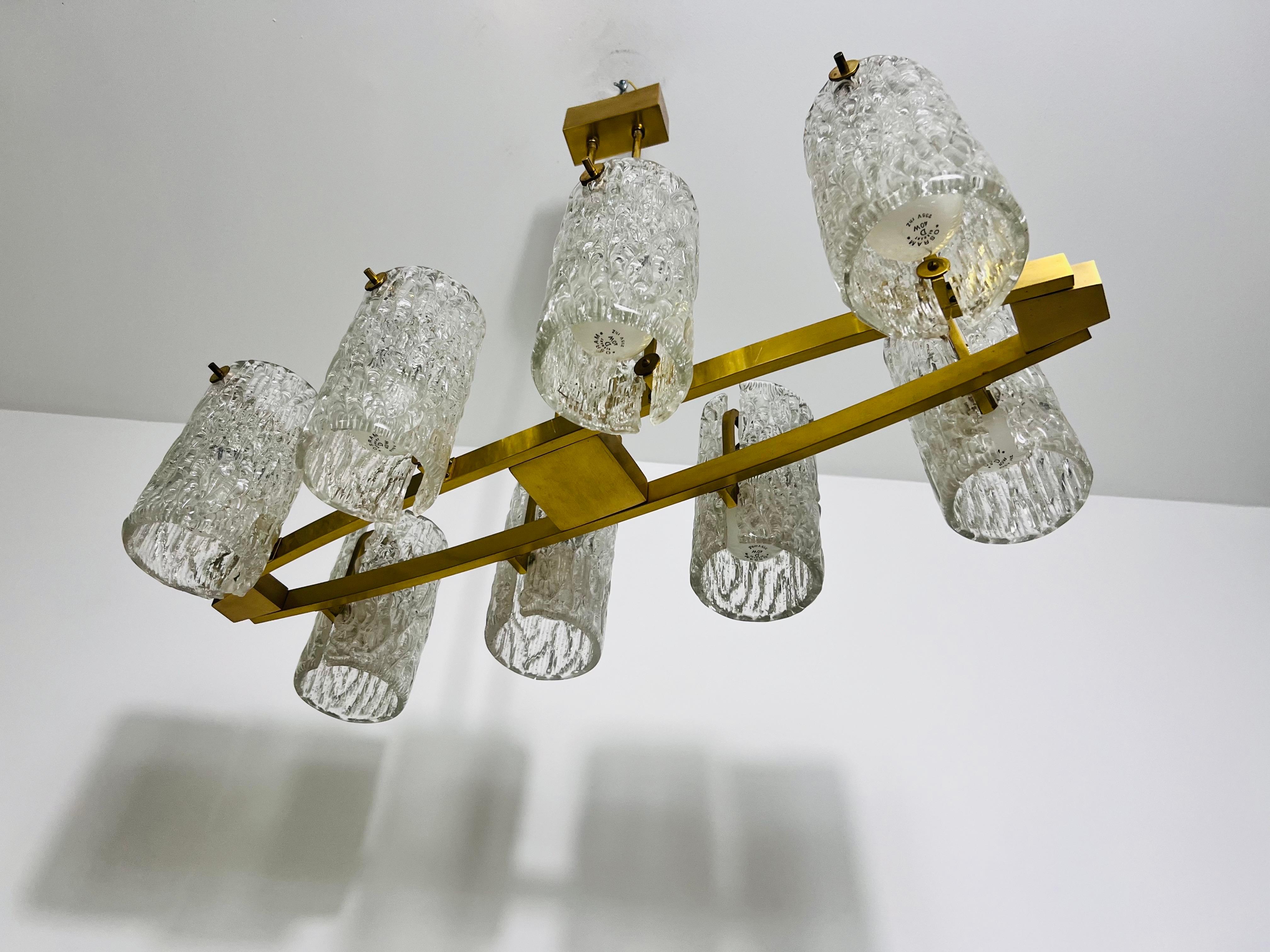 Questo elegante lampadario in ottone degli anni '60 sfoggia uno splendido design Mid-Century Modern influenzato dal movimento Bauhaus. Il lampadario presenta otto bracci, ognuno dei quali è ornato da un paralume in vetro ghiaccio testurizzato, che