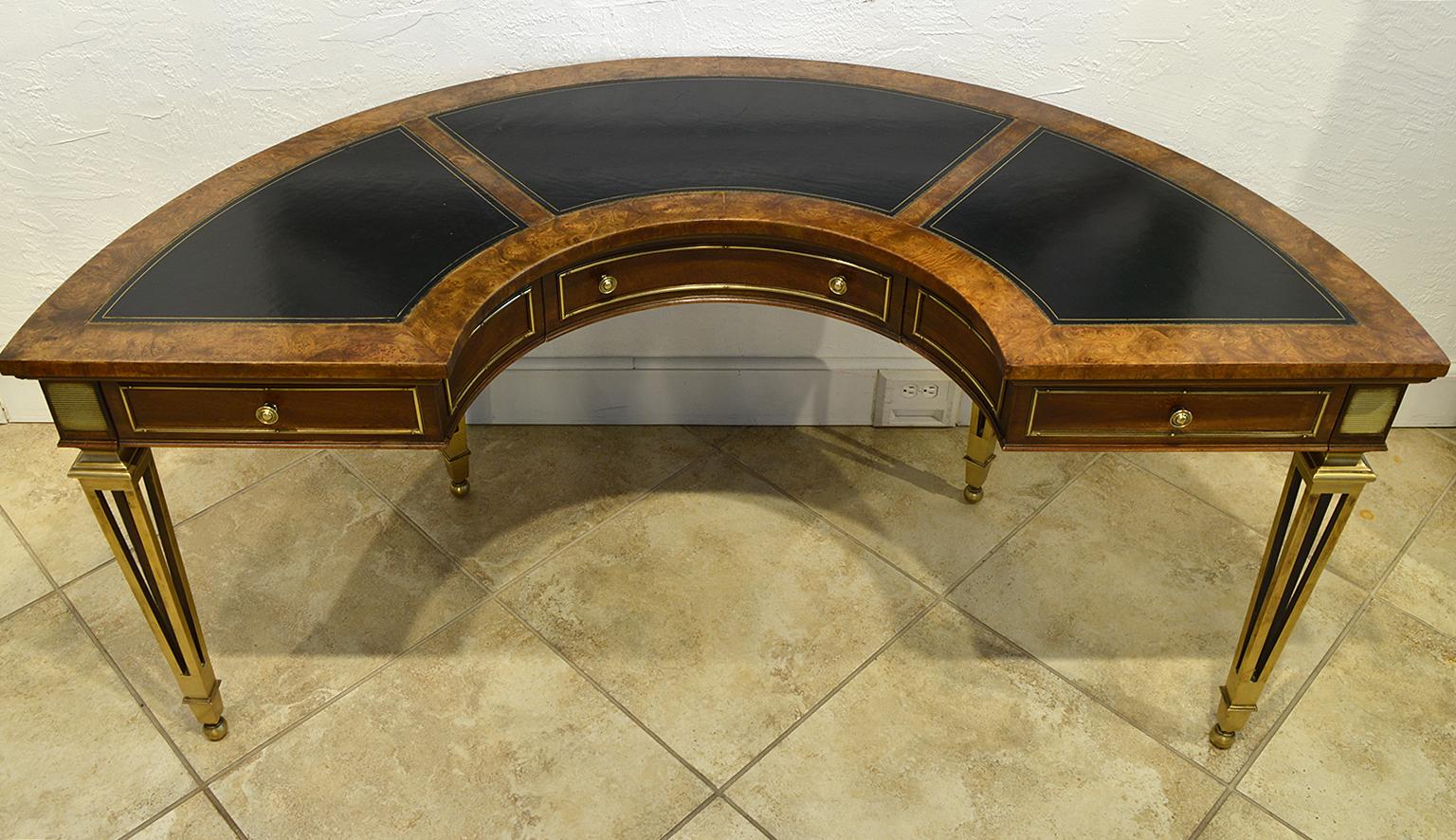 This magnificent Mastercraft semi-circular desk features a top inlaid with three panels of gilt tooled leather above three brass trimmed drawers and brass trimmed panels on the back. The desk is resting on four elegantly designed tapering brass legs