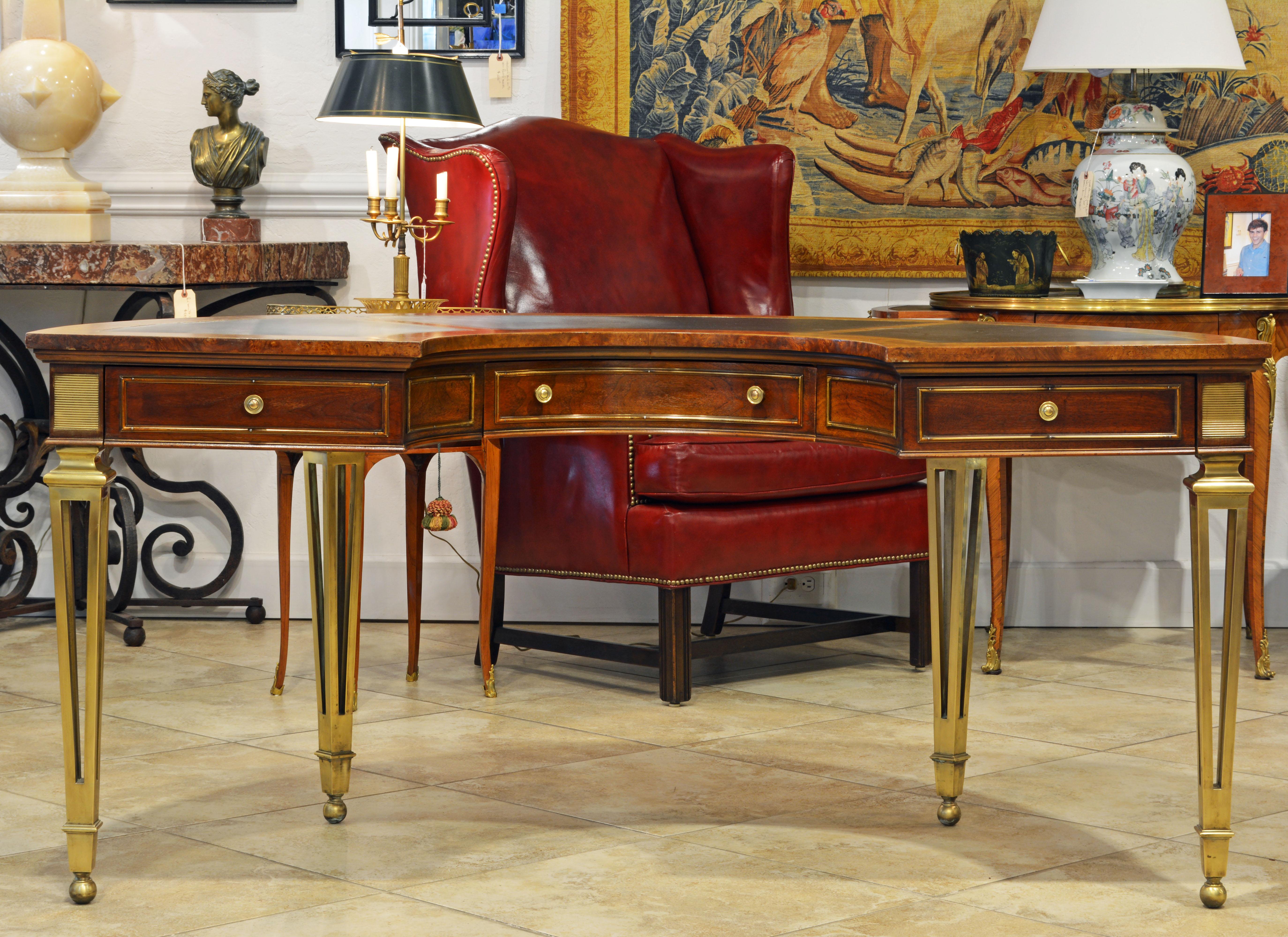 This magnificent mastercraft semi-circular desk features a top inlaid with three panels of gilt tooled leather above three brass trimmed drawers and brass trimmed panels on the back. The desk is resting on four elegantly designed tapering brass legs