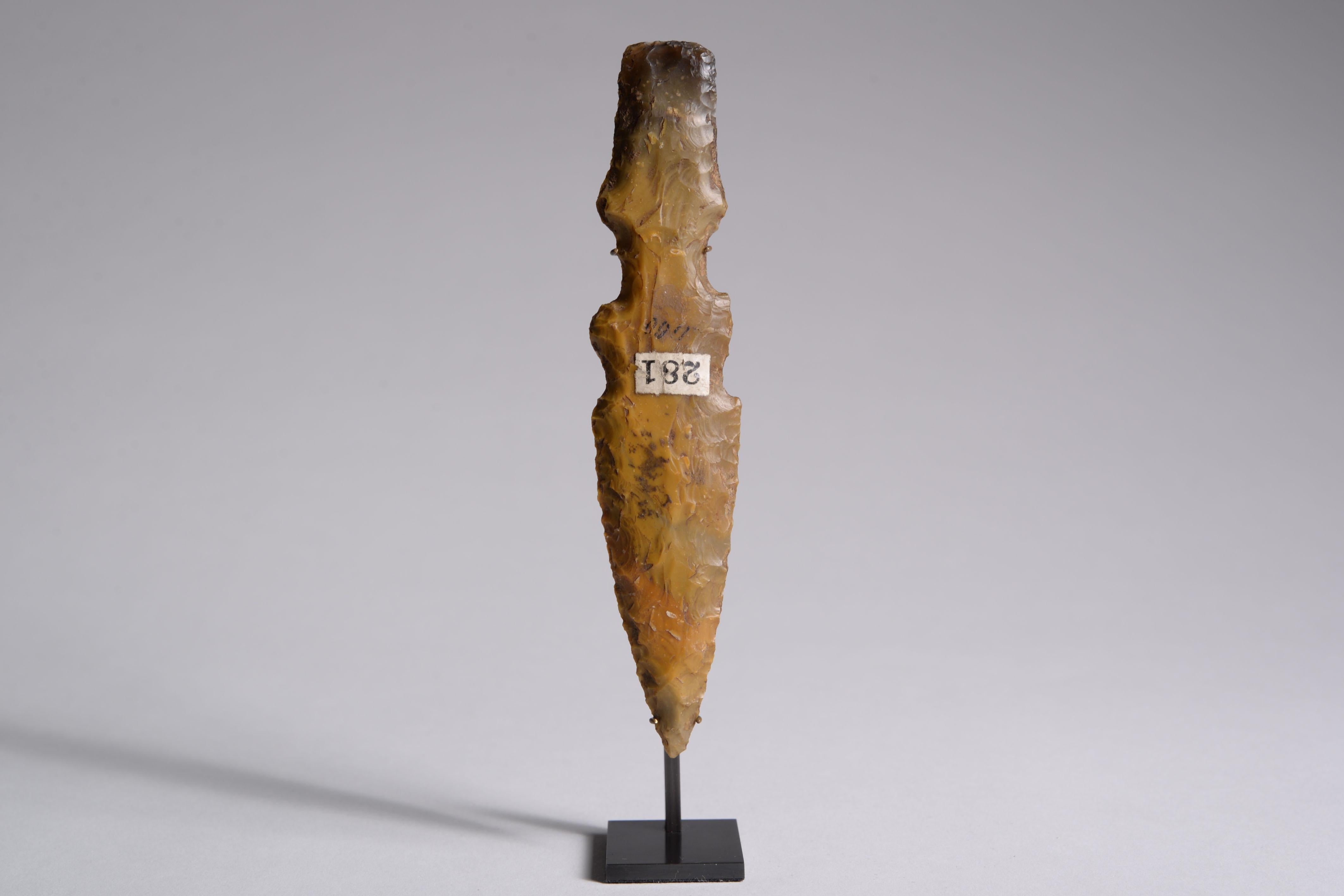 A beautifully modelled flint dagger, produced by the Bell Beaker culture: from the hilt to the tip, the stone fades from dark black to a burnt shade of yellow, creating a delicate interplay of colour across the face of the blade. The dagger is
