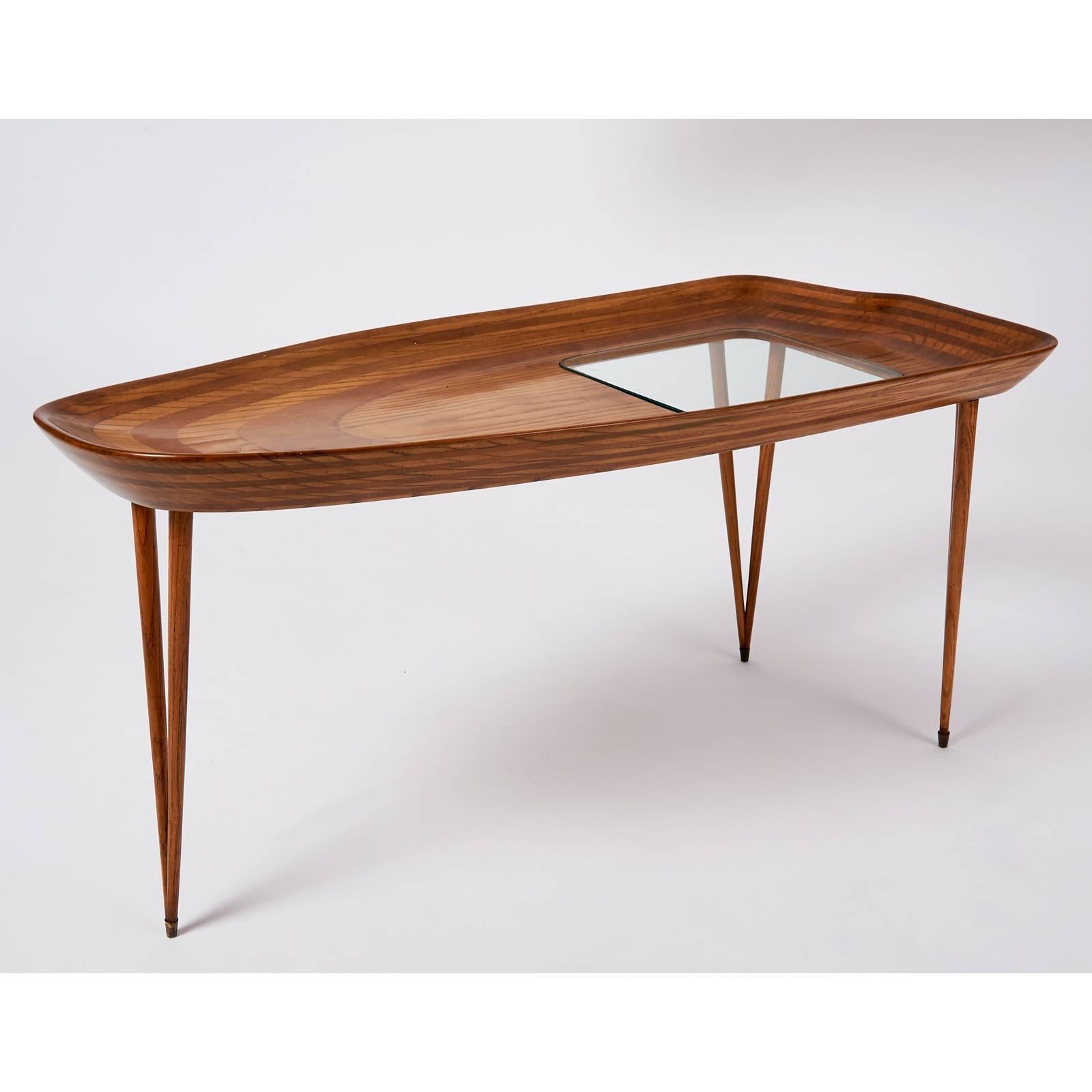 A beautifully formed oval table in laminated oak, organically shaped with turned up curving edges, sanded down to reveal the underlying layers of wood, with inset glass panel. Raised on three sharply tapered stiletto thin legs terminating in brass