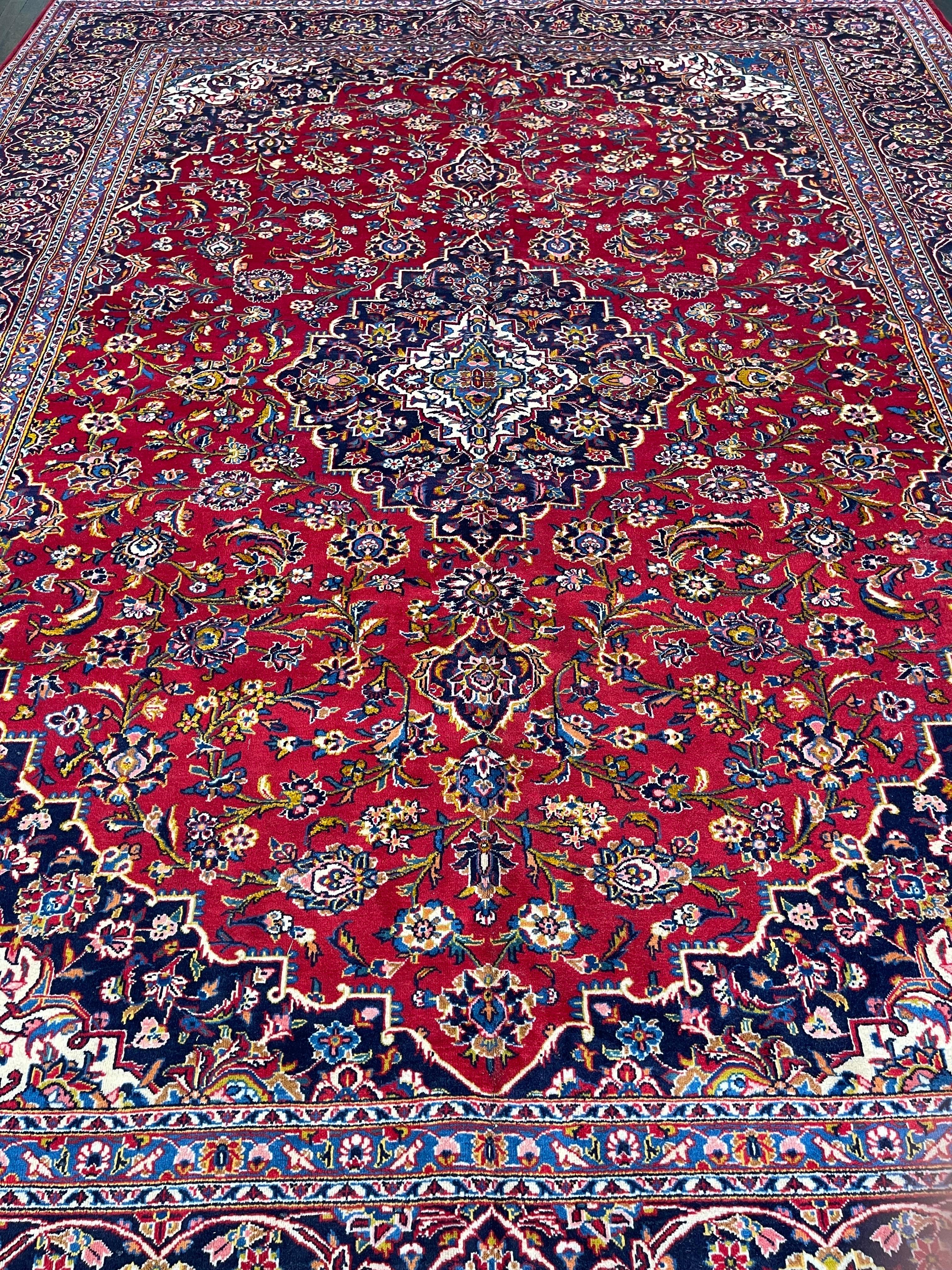 A fine persian carpet hand knotted in one of the major centers of rug making in Iran,this Kashan features a tomato red field decorated with flowers and leaves.The rug is designed with a navy blue medallion and surrounded with a indigo blue