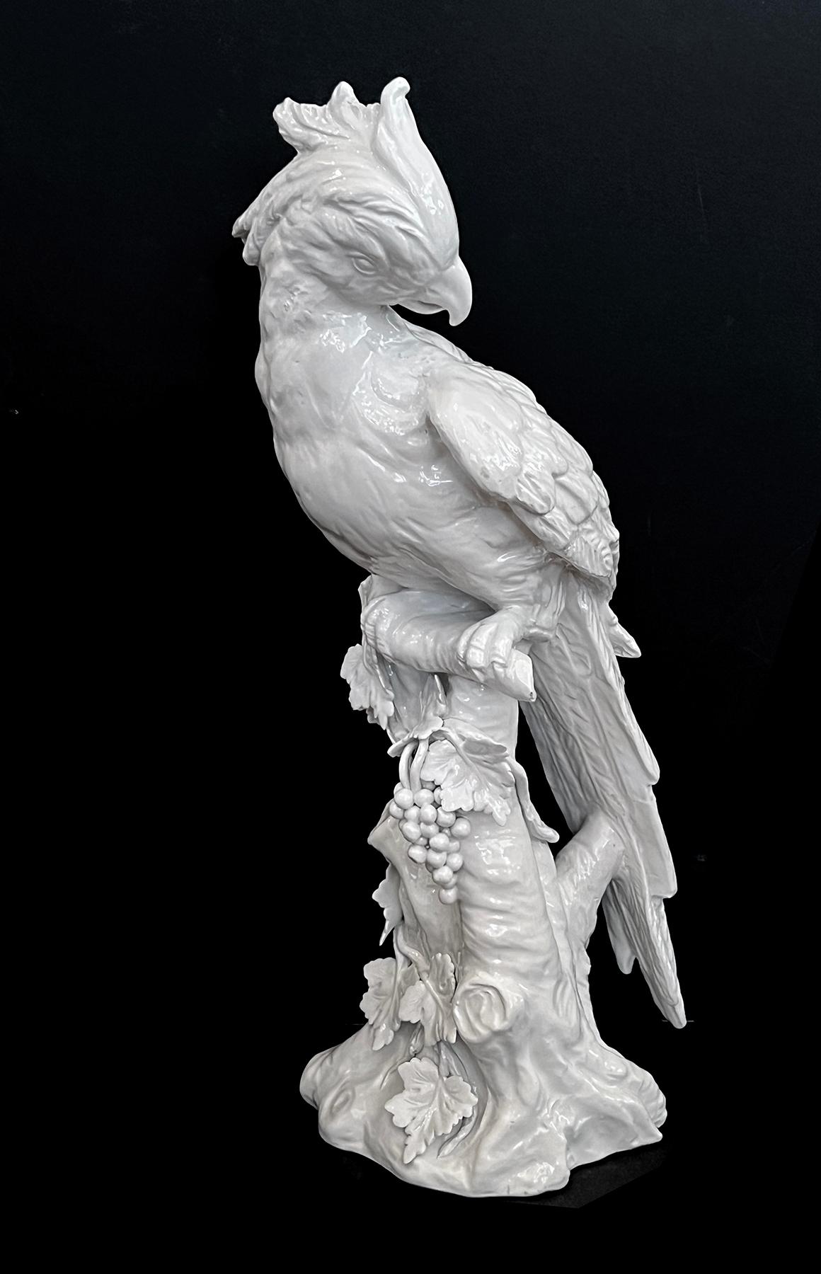 the large beautifully-detailed ceramic white glazed parrot with head in profile above a well-delineated plumed body with long tail feathers; all perched upon a branch-work base with applied leaves and grapes; with underglaze blue maker's mark and