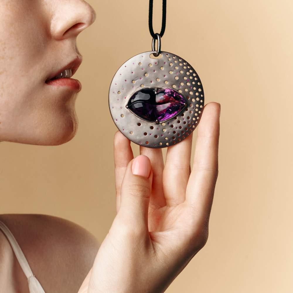 Smooth black rhodium battles with dots of color around a central amethyst gemstone. The small glimmers of ruby create a futher depth of color as they marry together on the surface of this shimmering pendant. The circular design creates a truly