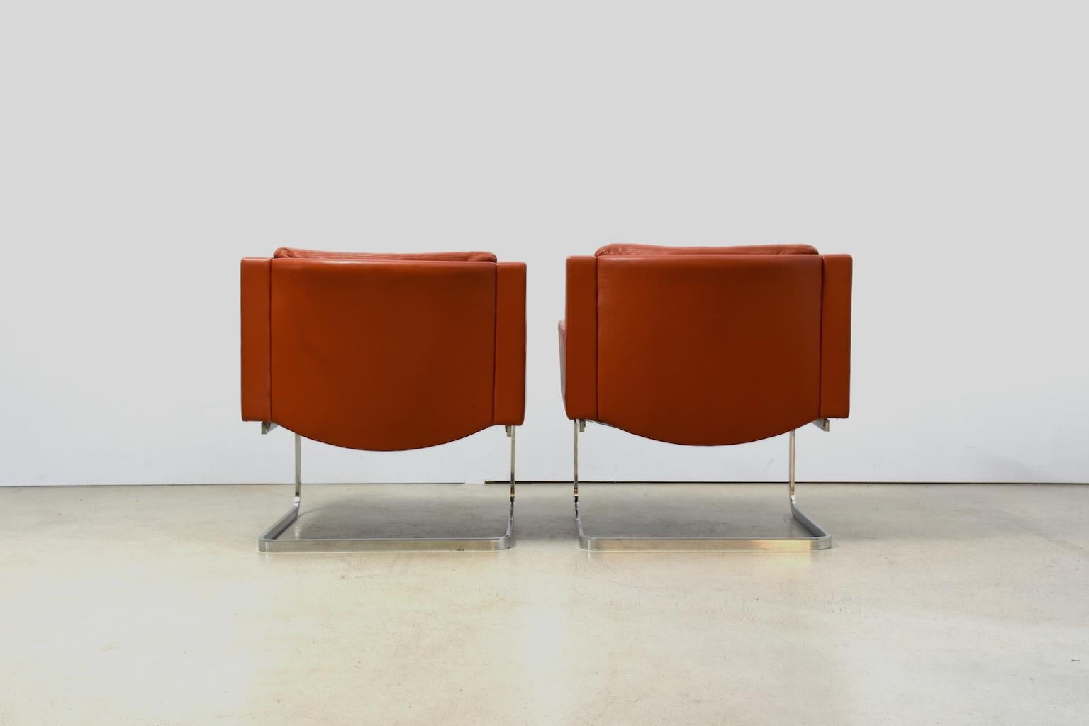 A pair of executive lounge chairs by Swiss architect and designer Robert Haussmann. Cantilevered construction with frame in chromed flat steel, body with wooden frame and covers in light reddish brown leather, leather cushions on the underside with