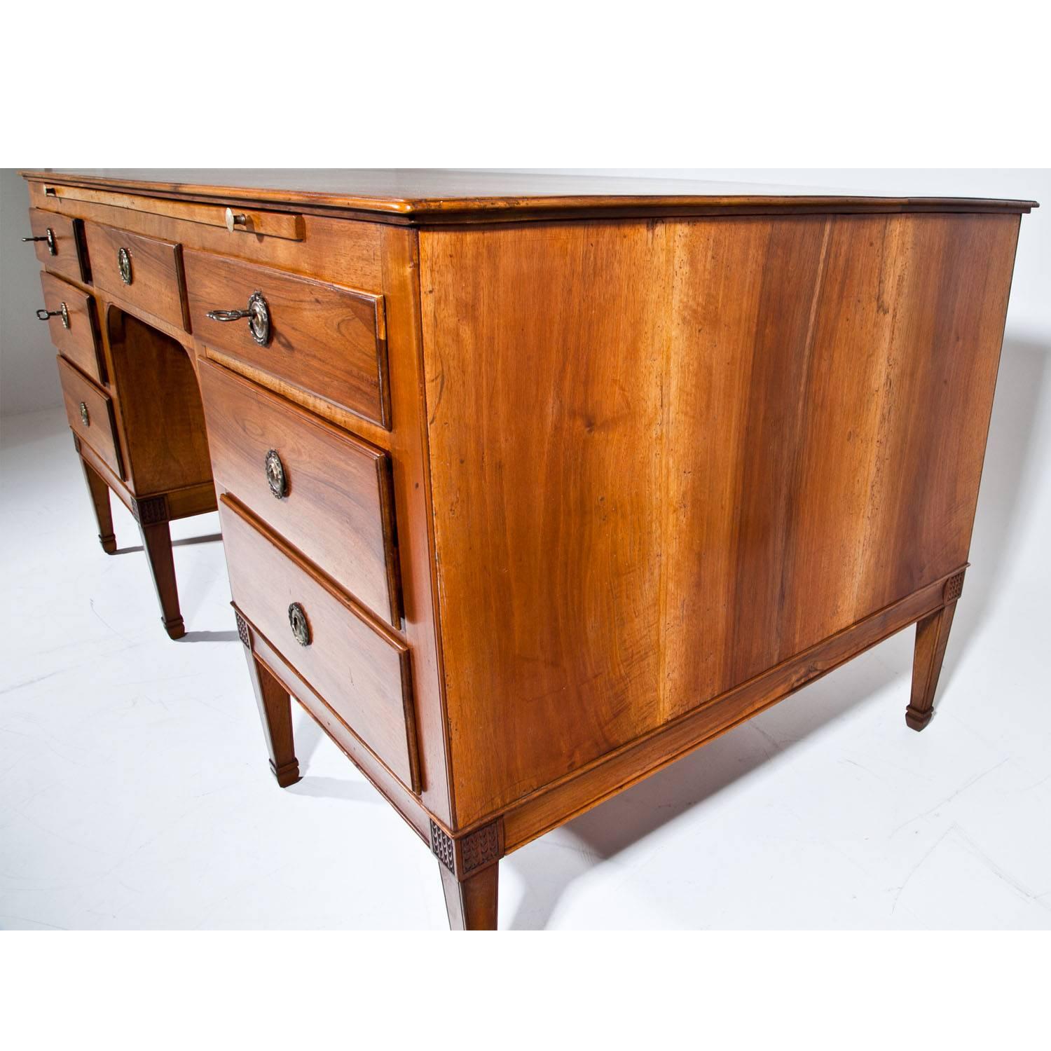 Large executive desk on eight tapered feet with small bases and a carved scale pattern. The desk has seven drawers and the back is designed in correspondence to the front, where fillings with brass fittings simulate drawers. A retractable writing