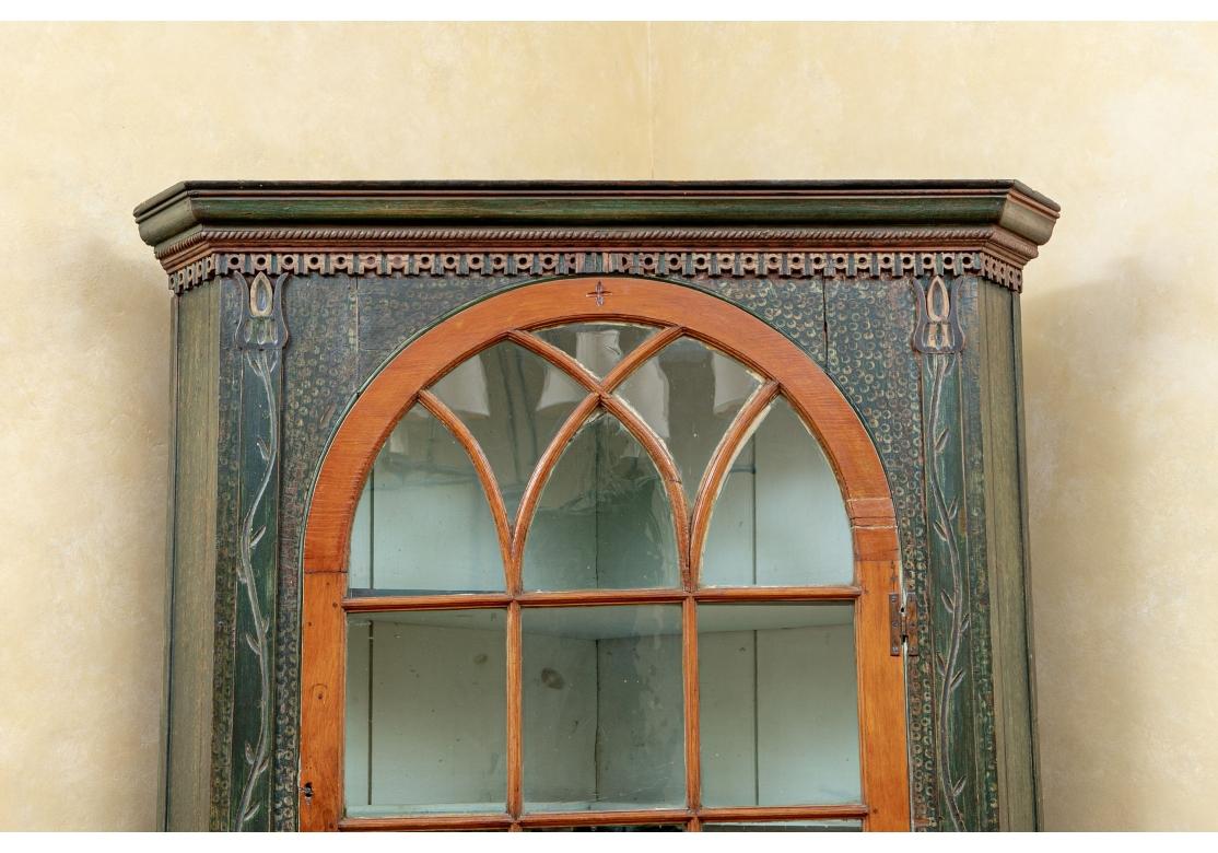With a Christies, New York auction sticker on the back. The cabinet with carved cornice and openwork frieze. Carved sinuous floral vine supports flank the glass door with mullions in a light brown stain. The interior in pale green paint with three