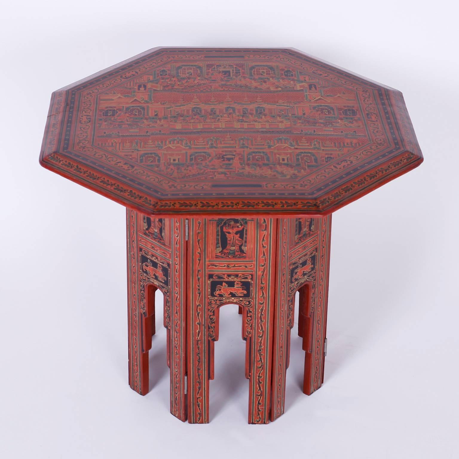 Exotic Thai table with an octagon top hand-painted with elaborate temple scenes and floral borders over a cinnabar colored background. The eight sided arched base is decorated all around with cats and flowers.