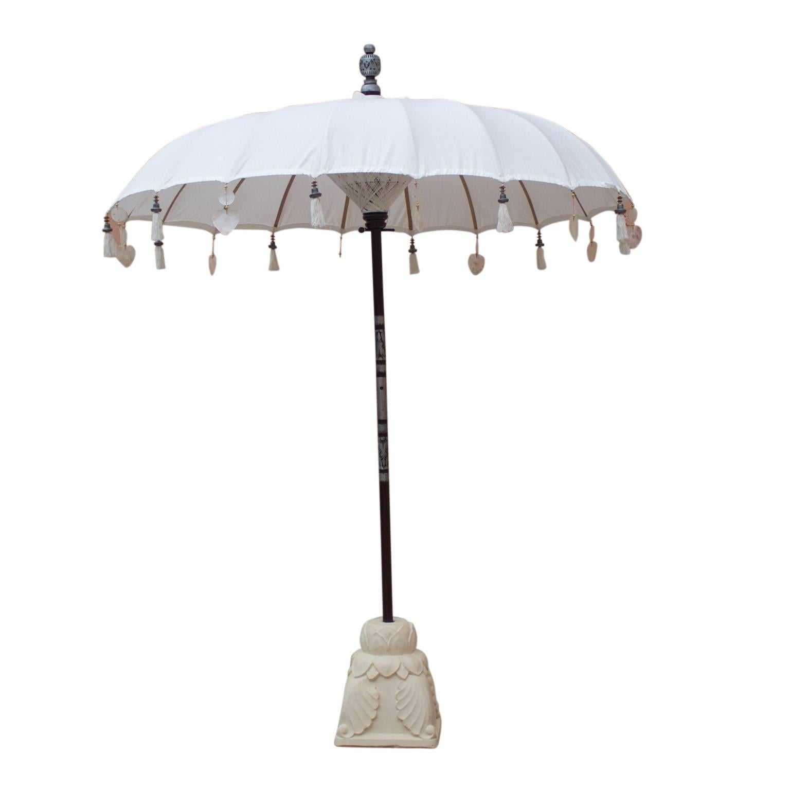 Exotic white cotton umbrella with bamboo ribs trimmed with carved wood and mother of pearl drops. The two-piece pole is decorated with tribal unscripted metal, as is the finial. All of this set in a heavy hand cast sandstone base with lotus and