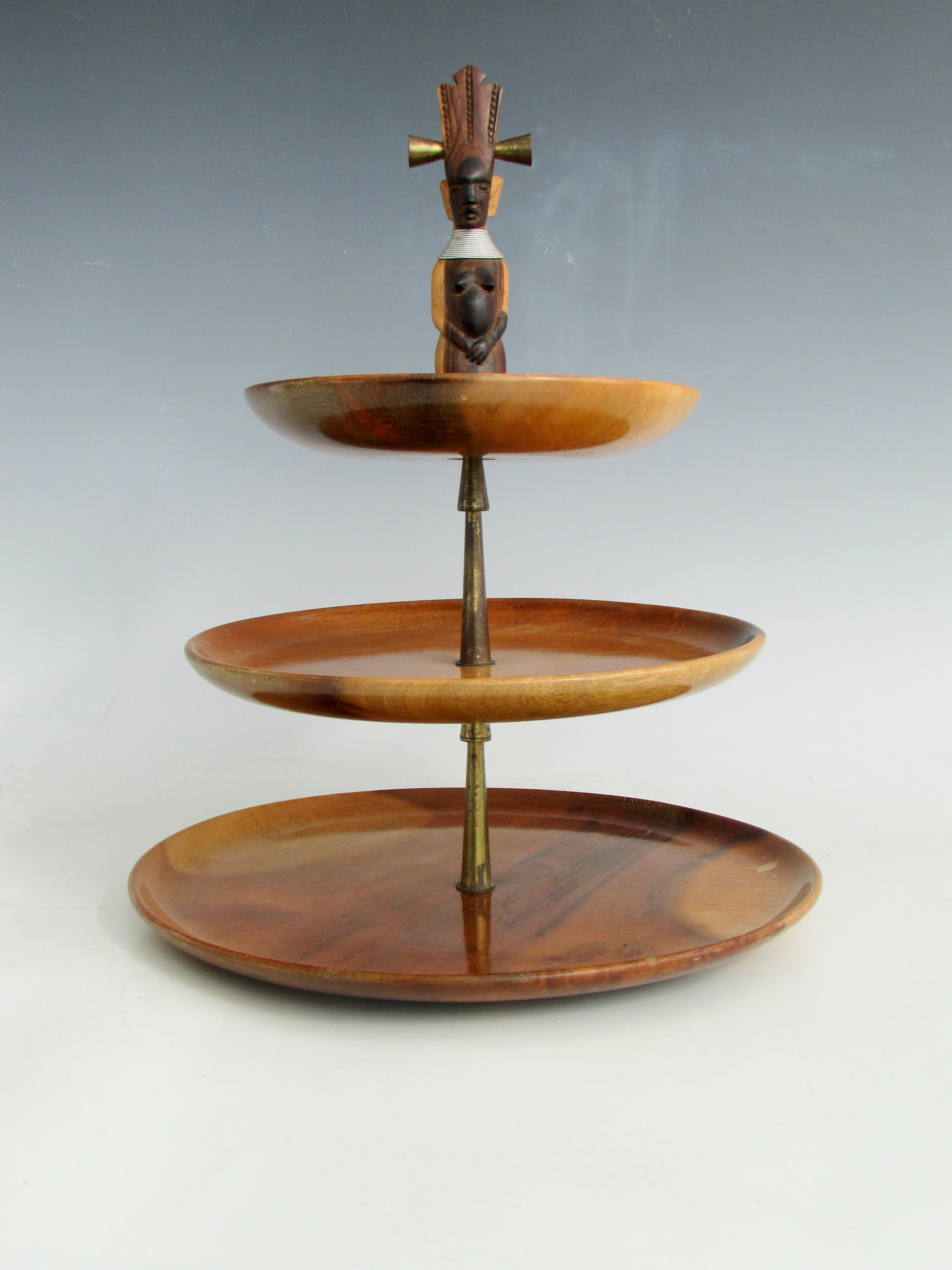 Three tier serving tray with carved exotic wood Tiki finial handle . Tiers taper largest on bottom getting progressively smaller . Center shaft is turned brass in an Art Deco stepped design . Fine original condition possibly never used .