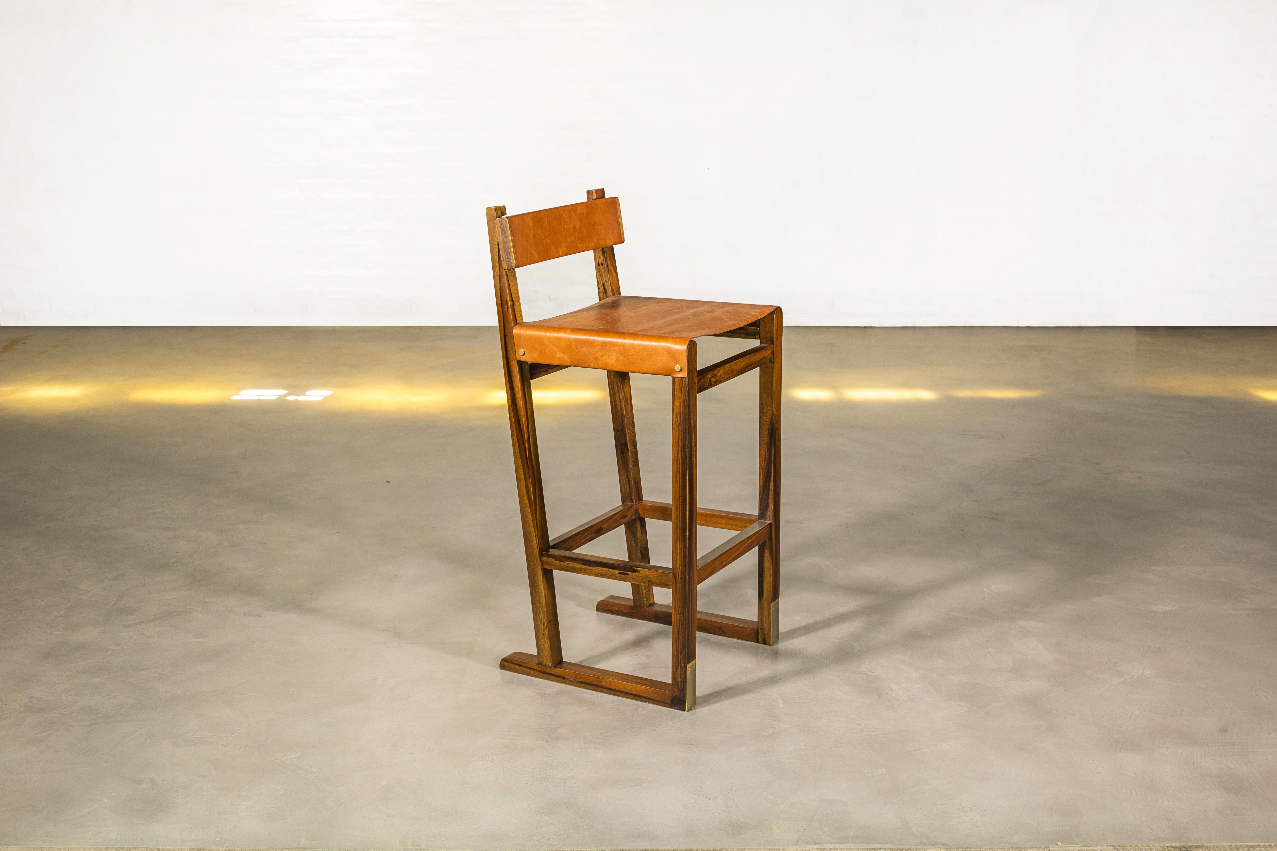 Le tabouret Piero présente une structure angulaire en bois massif avec une assise en cuir, un dossier en bois gainé de cuir et un repose-pieds en bronze bruni. Disponible dans n'importe quelle couleur de cuir ou dans le matériau d'ameublement de