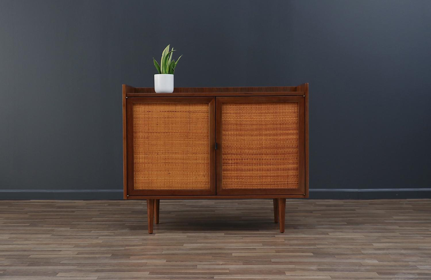 Enhance your home with the Jack Cartwright Cabinet with Cane Doors, crafted for Founders Furniture Company in the 1960s. Made from walnut wood, it features original cane front doors and a new leather pull. Newly refinished, it offers ample storage