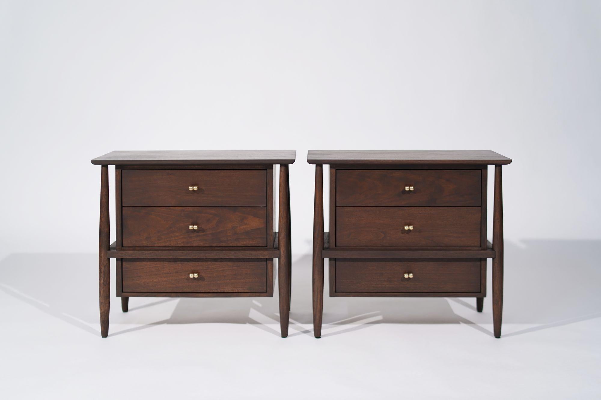 Pair of mid-century modern bedside tables or nightstands crafted in walnut by John Stuart, circa 1950-1950. These stunning pieces showcase an elegant exposed framework, three spacious drawers, and luxurious brass hardware, blending timeless design
