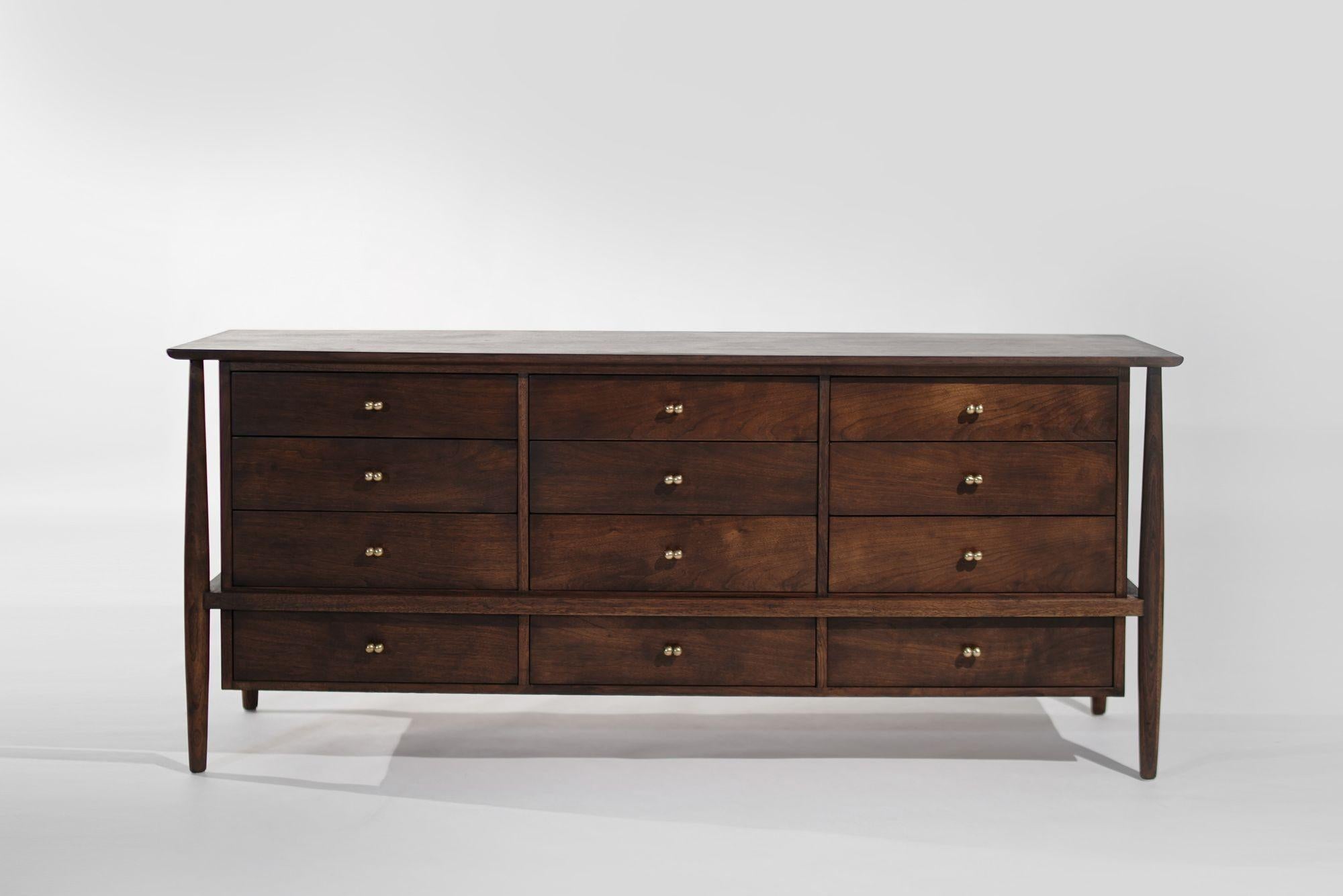 Mid-century Modern dresser in walnut by John Stuart, circa 1950-1950. This stunning piece showcases an elegant exposed framework, twelve spacious drawers, and luxurious brass hardware, blending timeless design with functional beauty.
