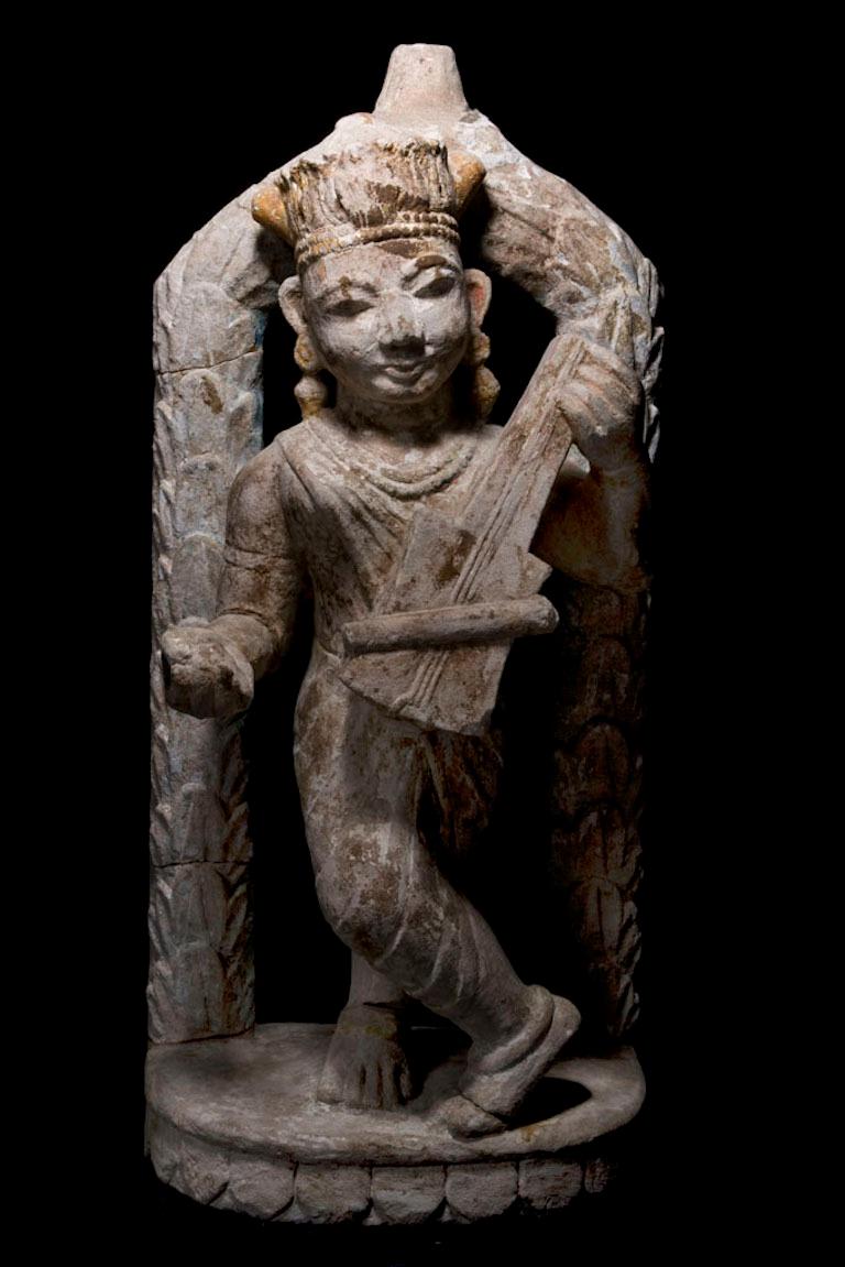 Sandstone sculpture of Apsara playing sitar with dancing pose in front of a garland arch. Covered with rests of stucco and old painting with beautiful face expression and flower lotus tiara. Gujarat style.