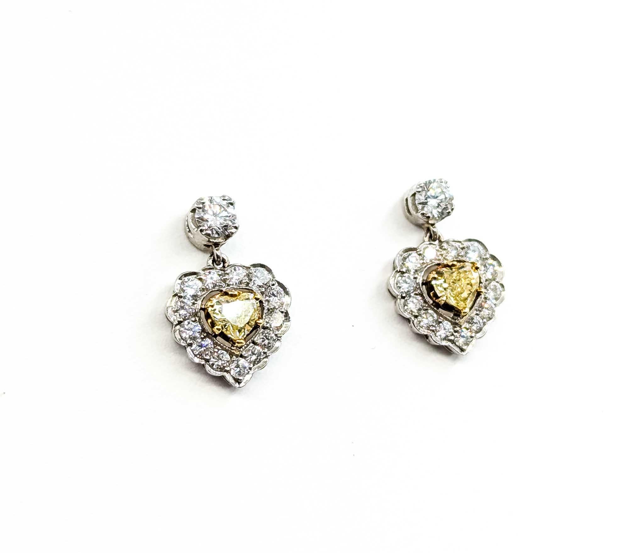 Estos impresionantes pendientes colgantes están elaborados en lujoso oro blanco de 18 quilates, diseñado para resaltar el brillo de los diamantes en talla redonda y corazón. En el centro de cada pendiente descansa un vibrante diamante amarillo en
