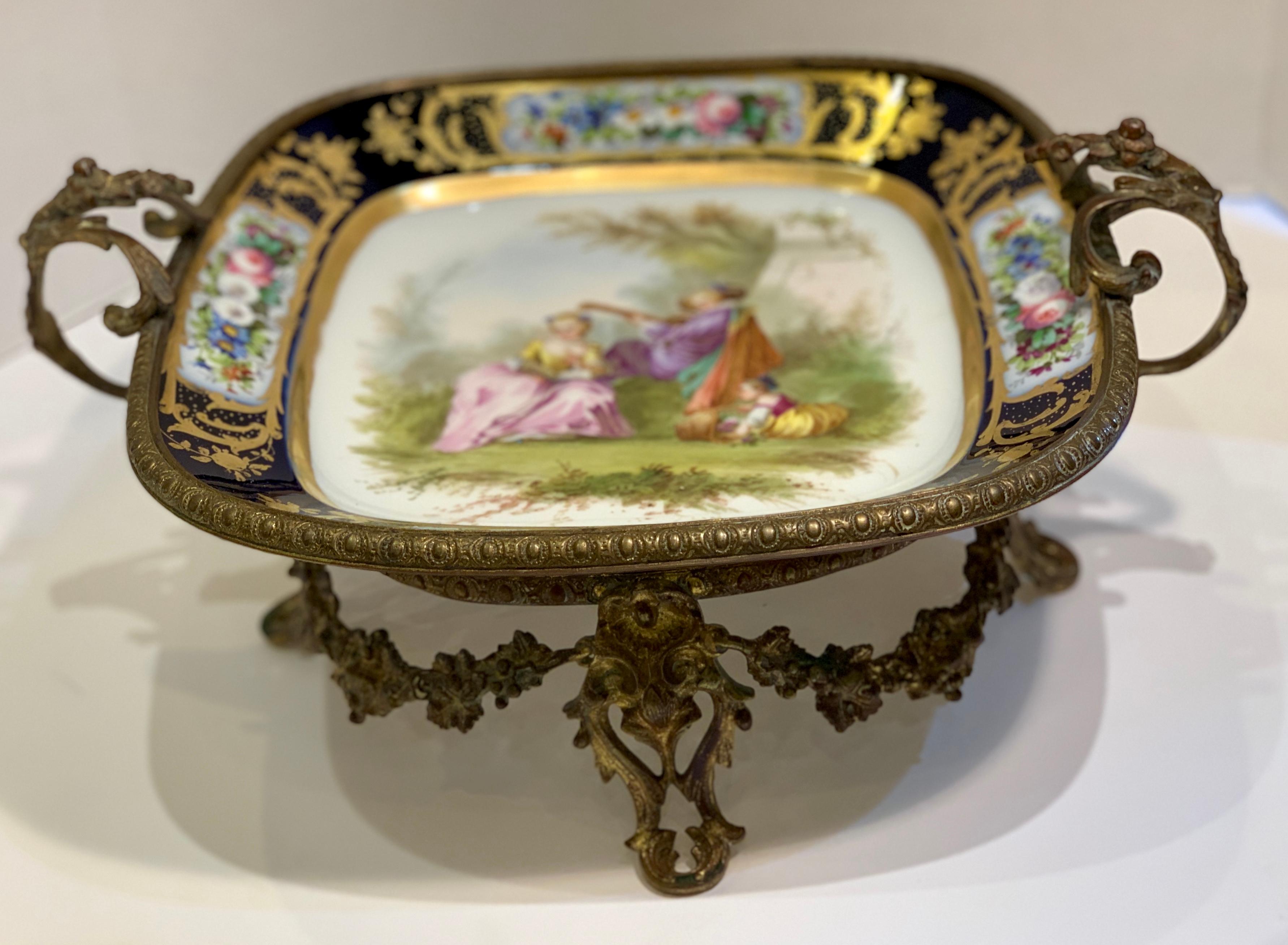 Opulent, 19th century handmade and painted square porcelain and ormolu gilt metal centerpiece features elaborately scrolling floral split handles, and a grapevine wreathed frame with egg and dart edges on cabriole legs. An idyllic pastoral scene is