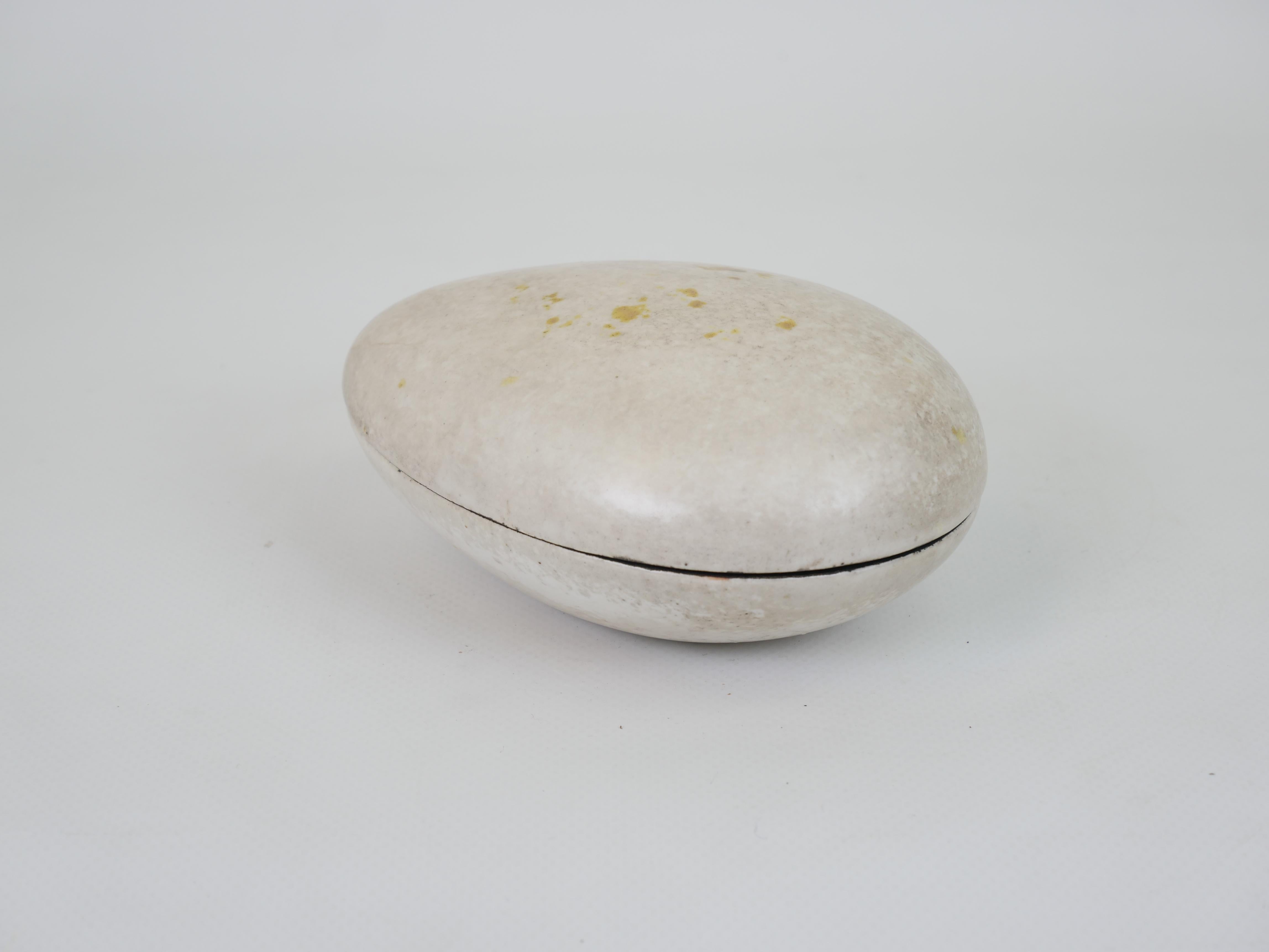 This exquisite ceramic box by renowned Swedish artist Hans Hedberg is crafted in the form of an organic, egg-shaped object. The exterior surface is smooth with a soft, natural beige glaze, delicately speckled with yellow accents, mimicking the