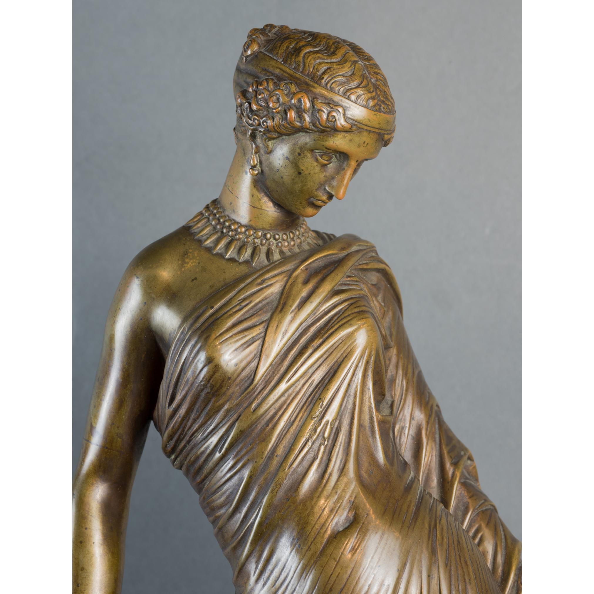 A beautiful female figural bronze statue of a lady wearing grecian style dress with thong sandals and an ornate necklace. She holds a Lyre which is carved in the pattern of a turtle shell. She leans on an ionic column that contains the signature,