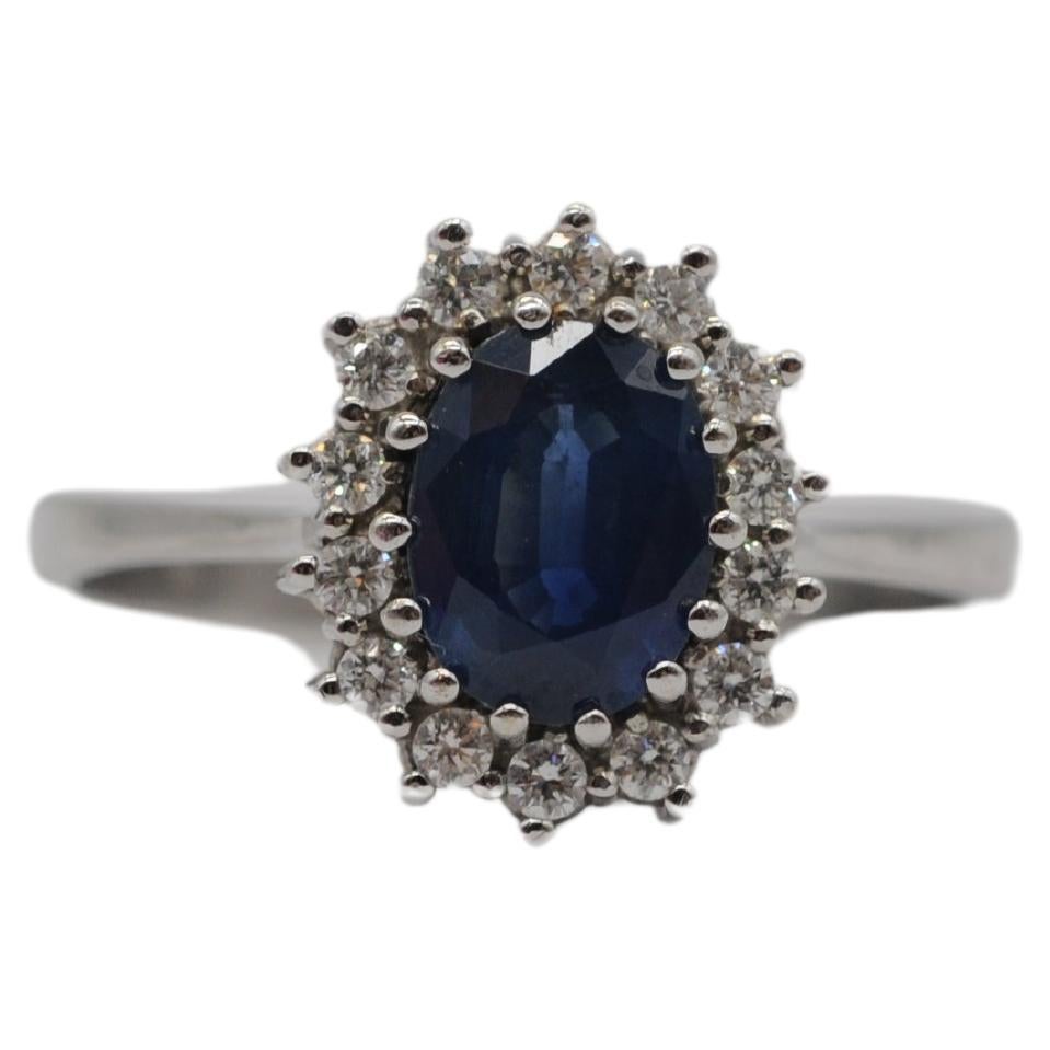 Immerse yourself in the regal elegance of this majestic 14k white gold ring, adorned with a captivating blue sapphire in an oval cut, weighing approximately 1 carat. Surrounding this exquisite centerpiece are fourteen delicate diamonds, each